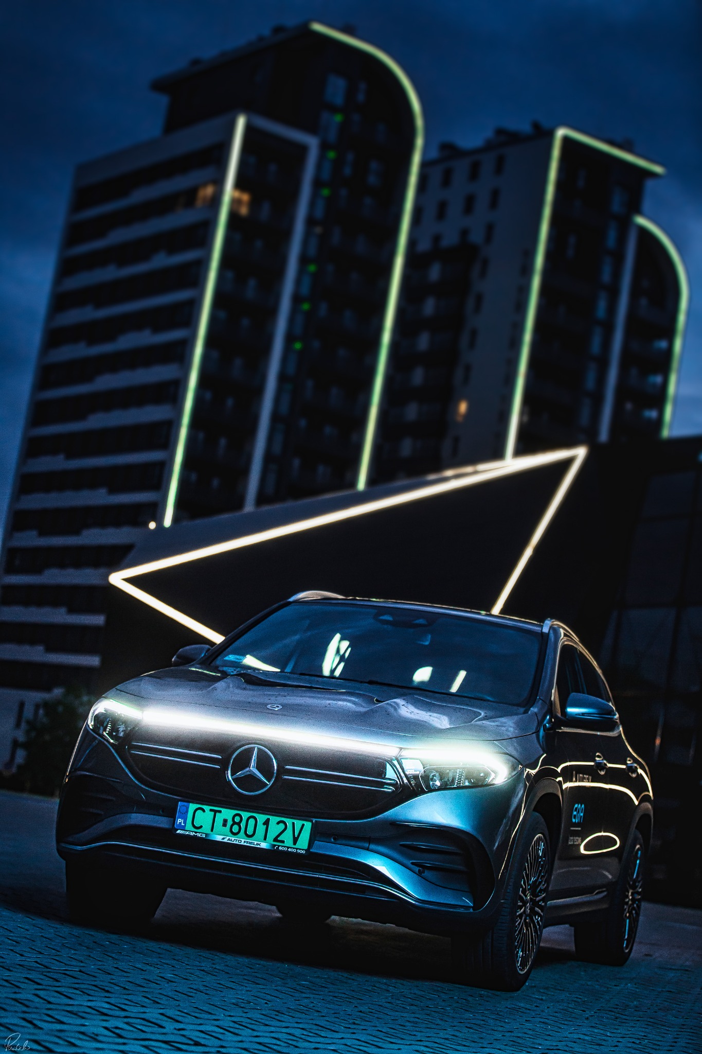 Nowoczesny, elektryczny samochód marki Mercedes-Benz EQA, sfotografowany nocą na tle budynku z neonowym oświetleniem, tablica rejestracyjna z zielonym tłem.