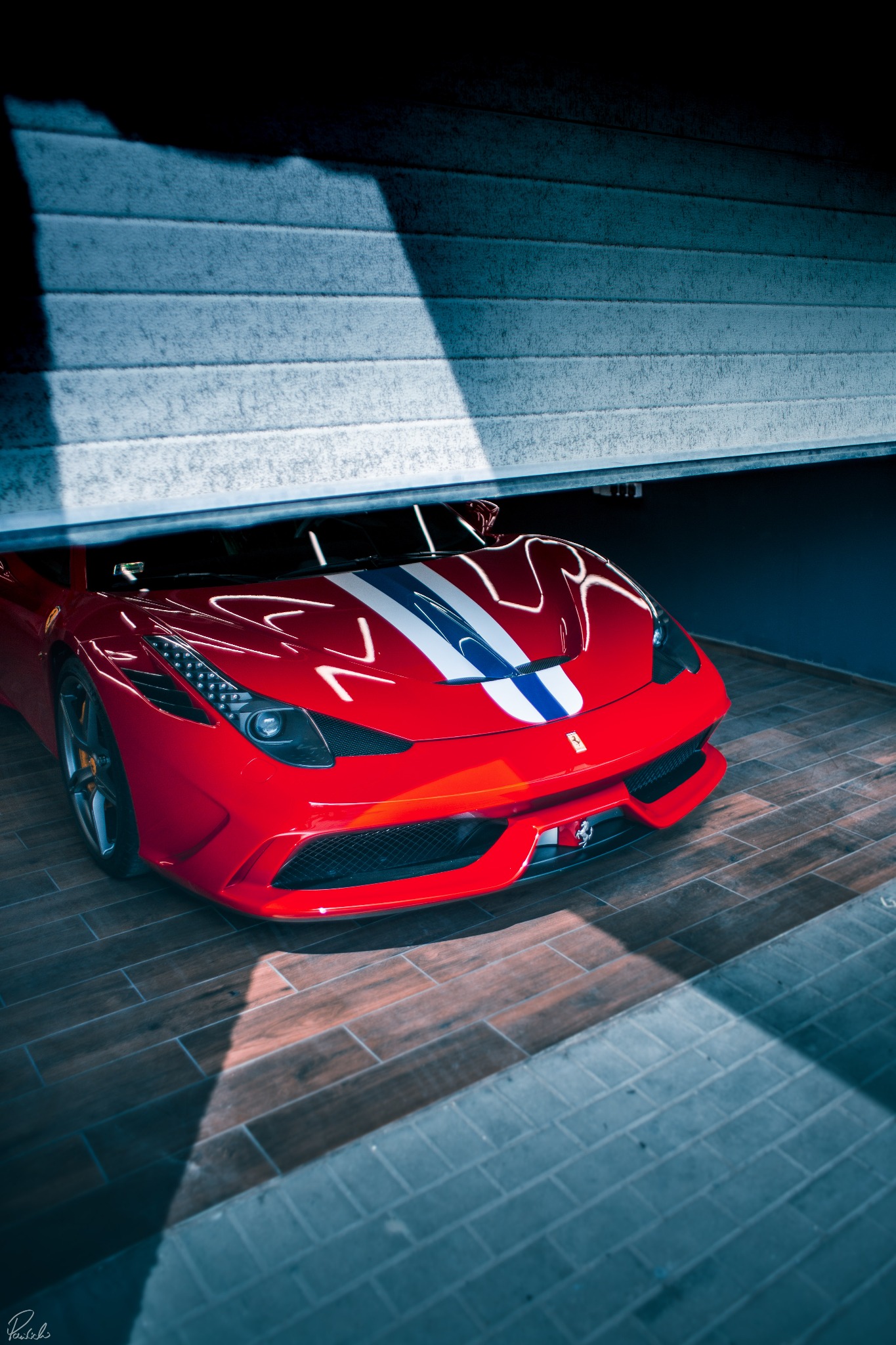 Czerwone Ferrari z białymi i niebieskimi pasami, częściowo schowane za uchyloną szarą bramą garażową, na drewnopodobnej podłodze, oświetlone padającym światłem.