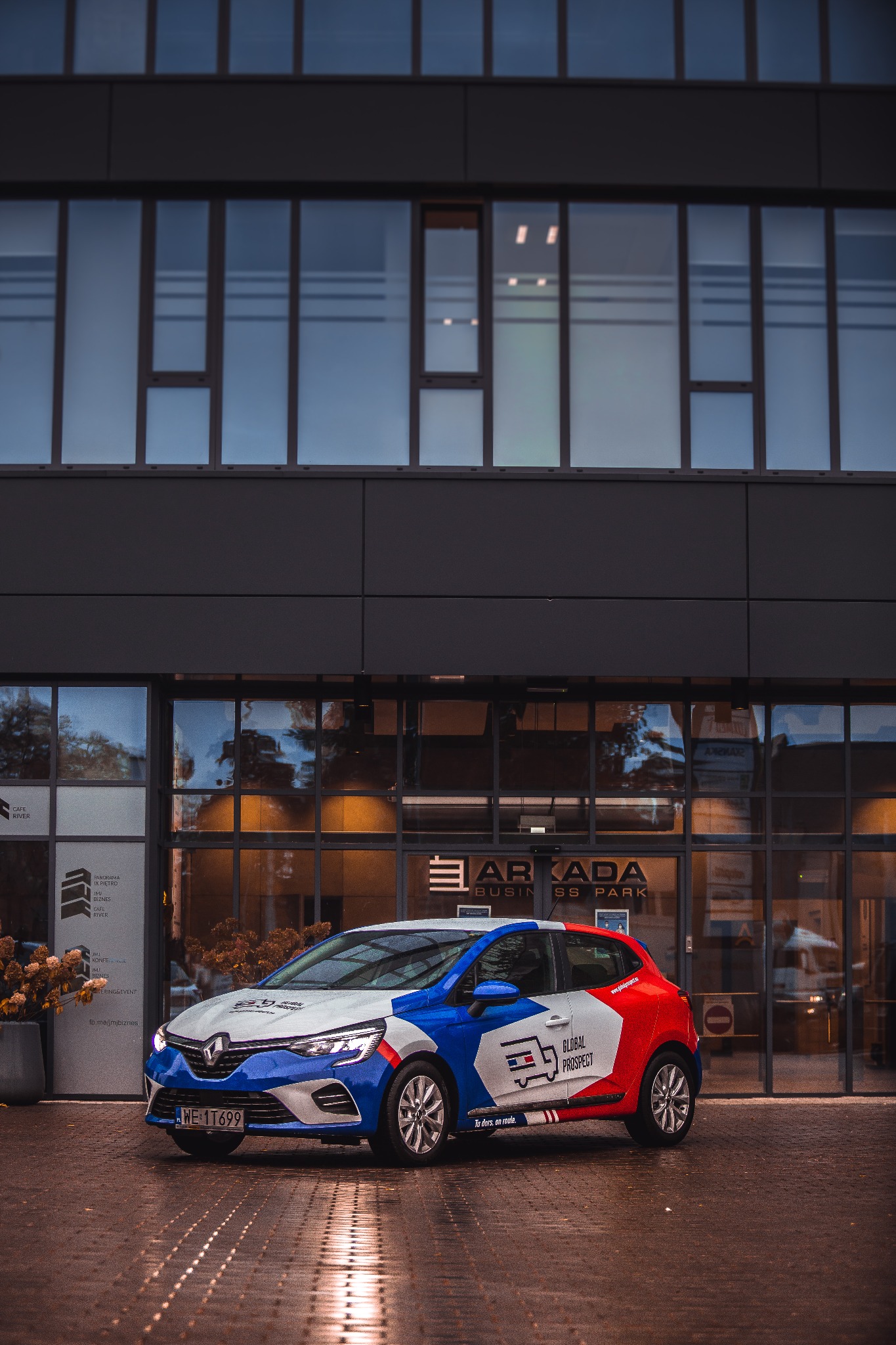 Samochód Renault Clio z brandingiem firmy Global Prospect zaparkowany przed budynkiem Arkada Business Park, widoczne odbicia w mokrej nawierzchni.