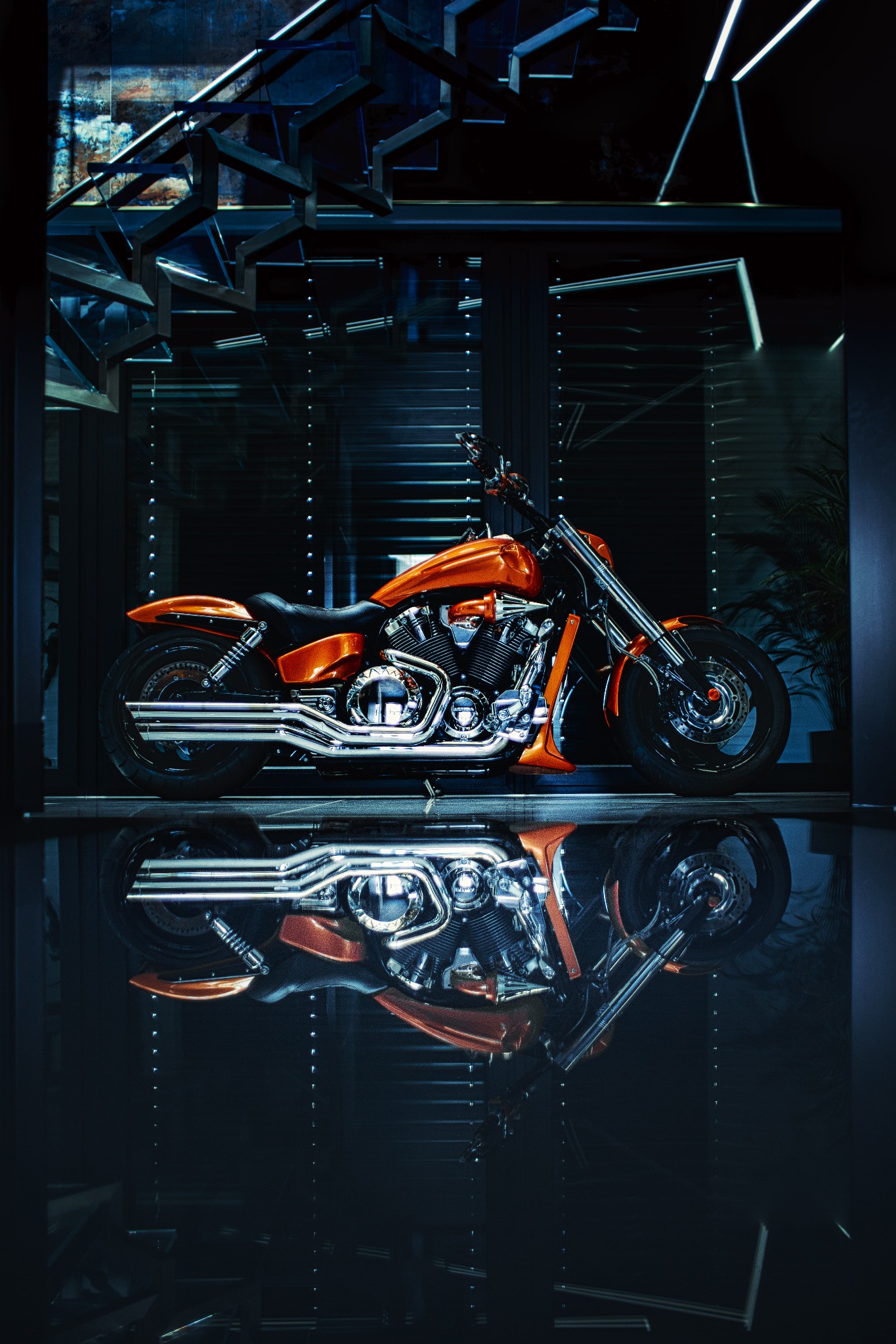 Błyszczący pomarańczowy motocykl typu cruiser zaparkowany w nowoczesnym wnętrzu z ciemnym, lustrzanym podłożem, odbijającym sylwetkę pojazdu.