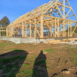 Surowy stan drewnianej konstrukcji domu na fundamencie z bloczków betonowych, widok na szkielet dachu i ścian w słoneczny dzień, cień fotografa w kadrze.