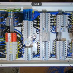 ELEKTRA Dariusz Glazer - Otwarta skrzynka elektryczna z widocznymi bezpiecznikami, przewodami i listwami zaciskowymi, częściowo zasłonięta dłonią z obrączką.