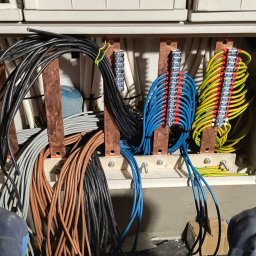 ELEKTRA Dariusz Glazer - Otwarta skrzynka rozdzielcza z licznymi przewodami w różnych kolorach (brązowe, czarne, szare, niebieskie, żółto-zielone) podłączonymi do listew zaciskowych na miedzianych szynach, widoczne...