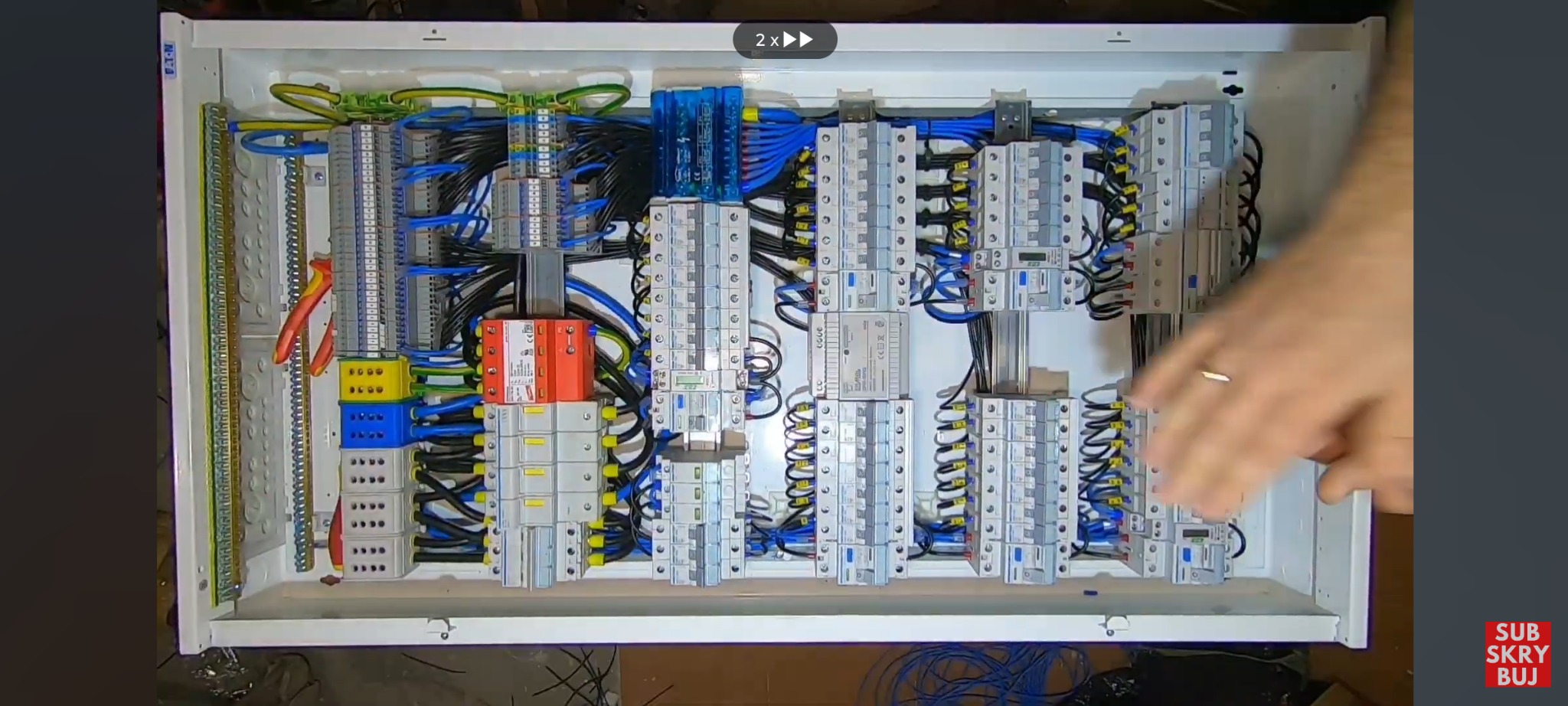 Otwarta skrzynka elektryczna z widocznymi bezpiecznikami, przewodami i listwami zaciskowymi, częściowo zasłonięta dłonią z obrączką.