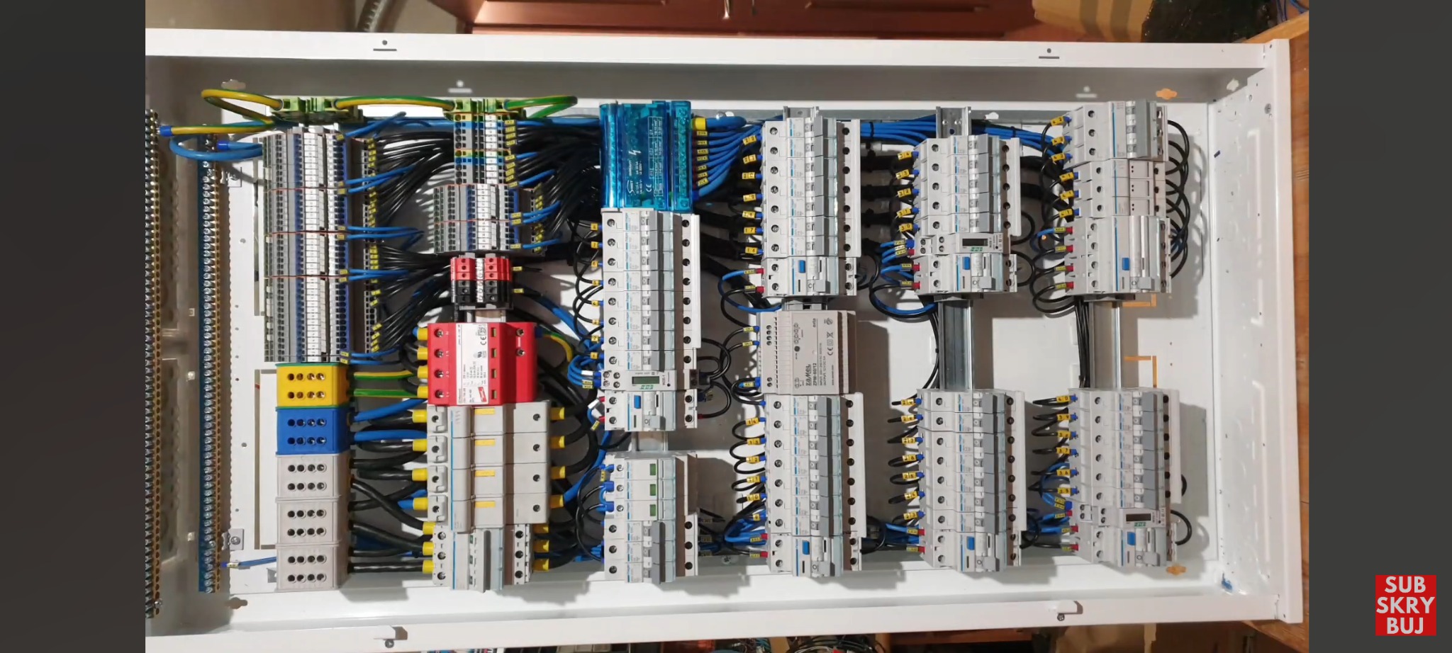 Rozdzielnia elektryczna w białej obudowie z widocznymi bezpiecznikami, przewodami w kolorach żółtym, niebieskim i czarnym, oraz listwami zaciskowymi, zainstalowana wewnątrz budynku.