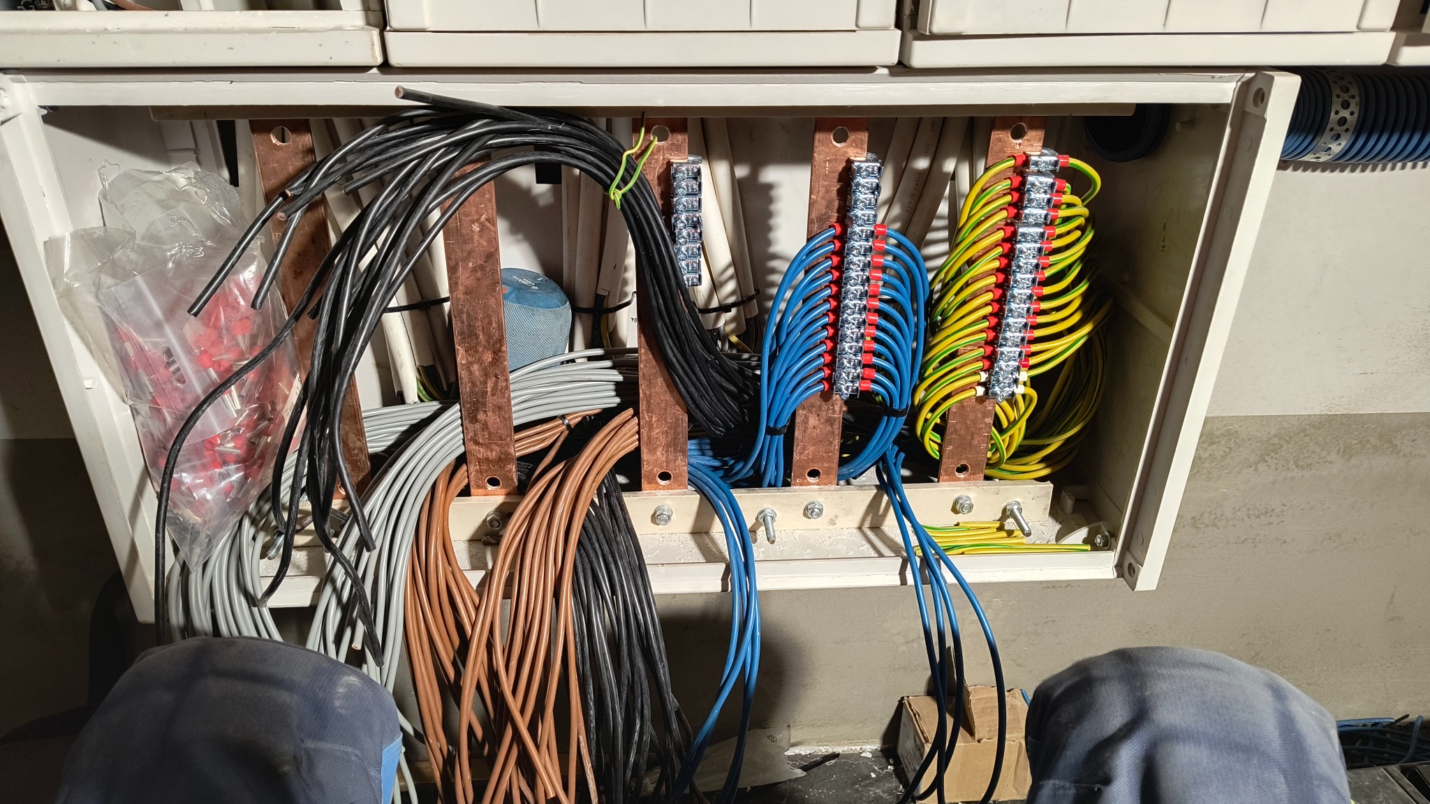 Otwarta skrzynka rozdzielcza z licznymi przewodami w różnych kolorach (brązowe, czarne, szare, niebieskie, żółto-zielone) podłączonymi do listew zaciskowych na miedzianych szynach, widoczne...