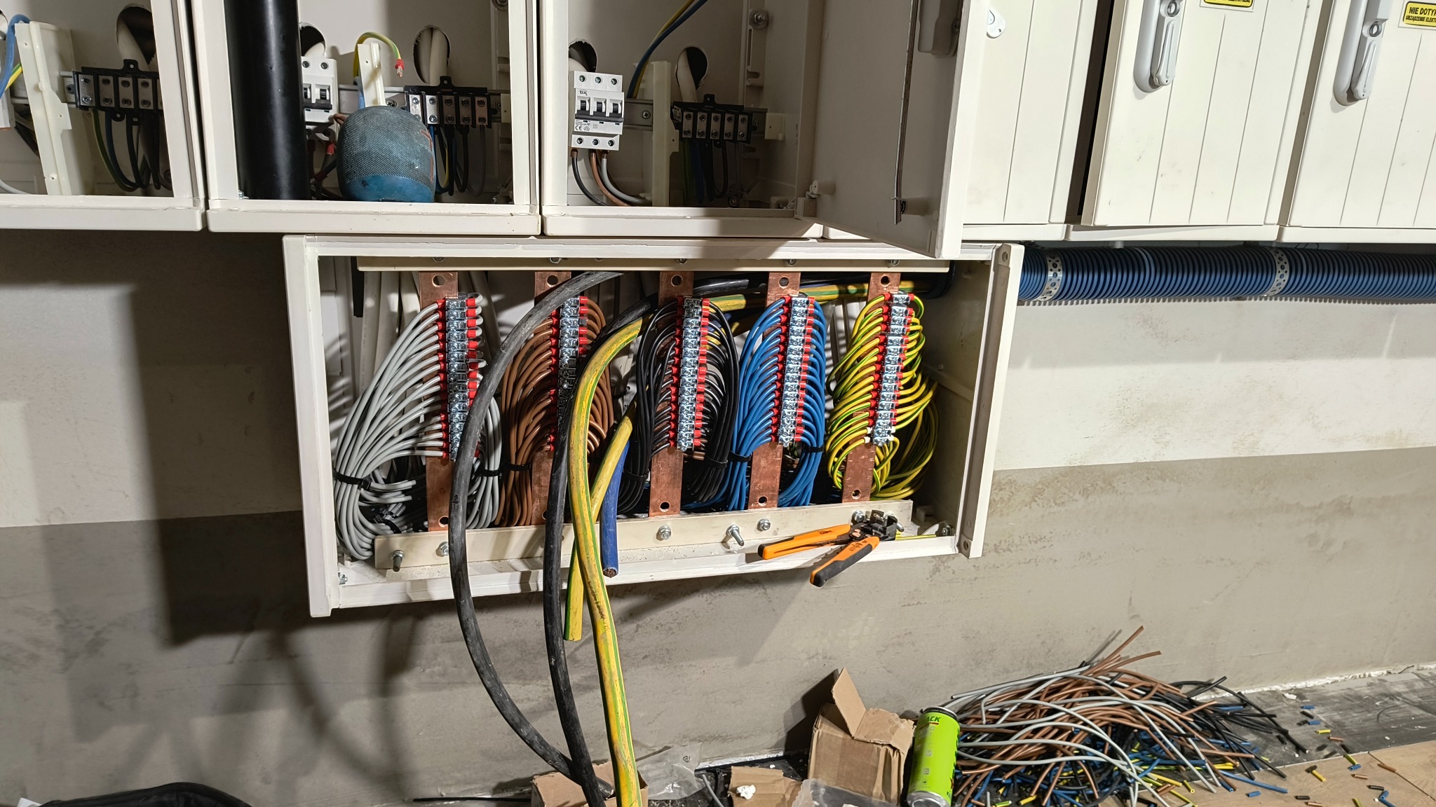 Rozdzielnica elektryczna z uporządkowanymi przewodami w różnych kolorach, widoczne listwy zaciskowe, narzędzia do obróbki kabli i fragment ściany w tle.