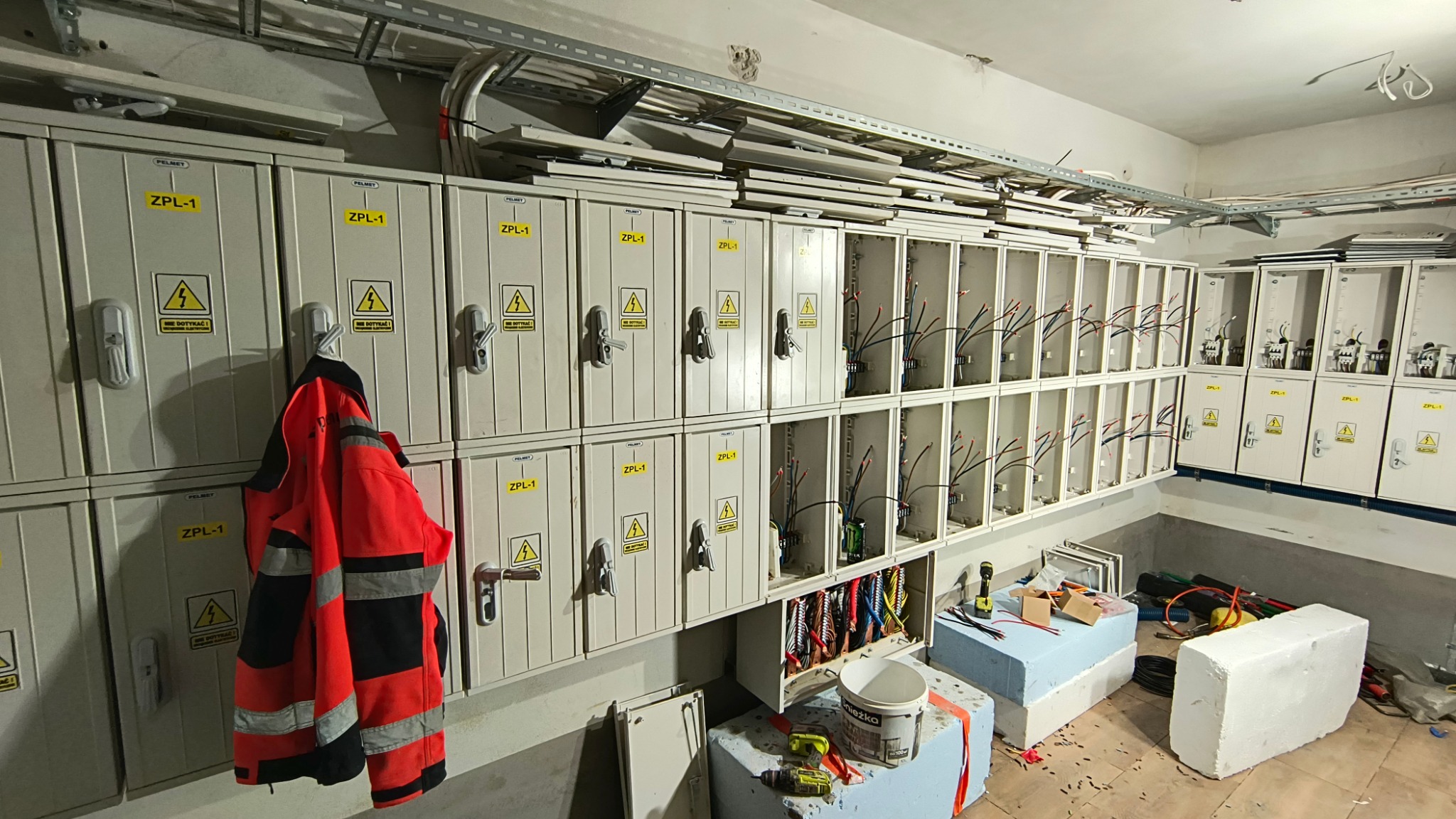 Szafy elektryczne w trakcie montażu, widoczne kable i narzędzia, na jednej z szaf wisi kurtka robocza.
