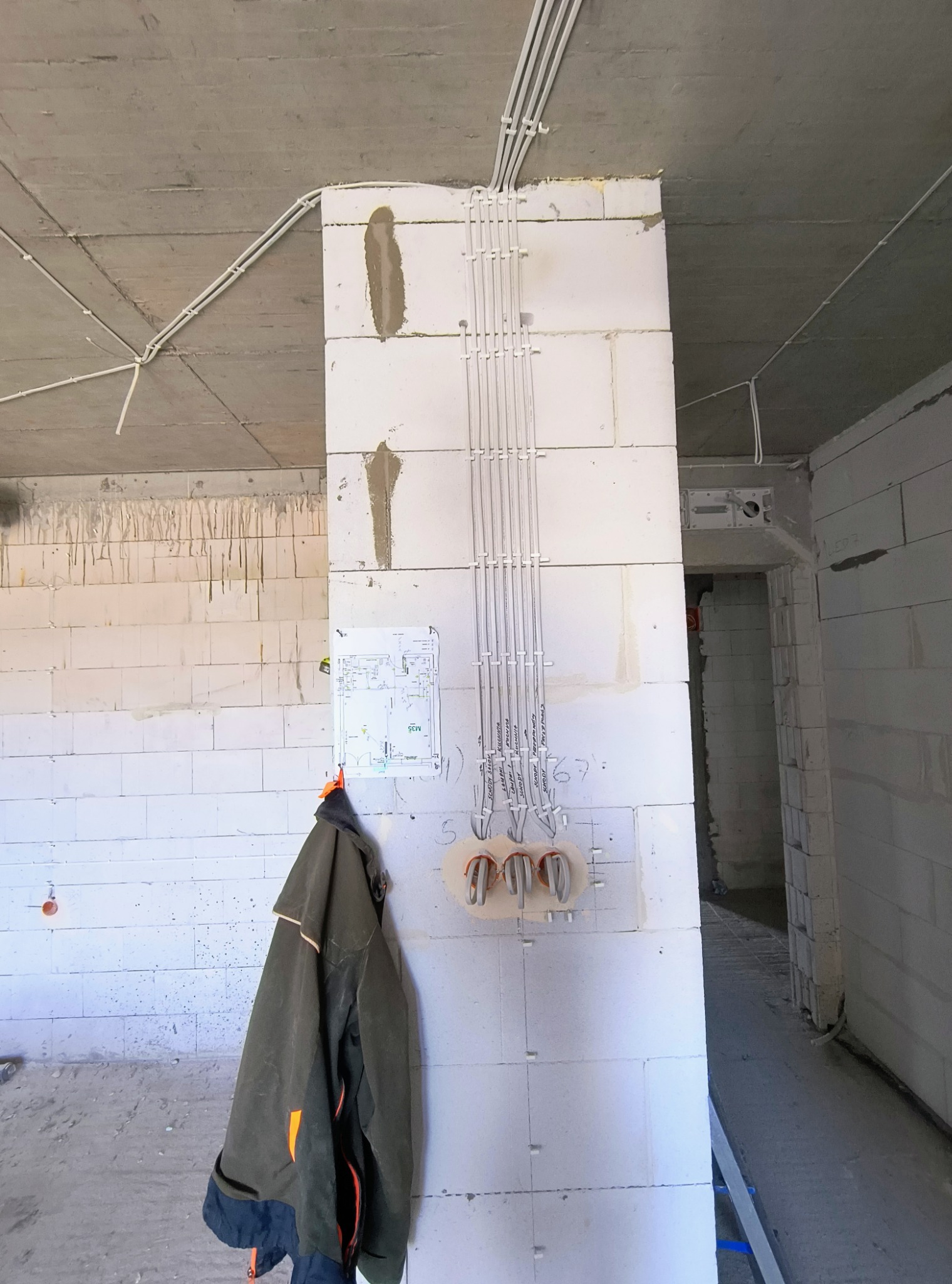 Wnętrze w trakcie budowy z widocznymi przewodami elektrycznymi prowadzonymi po ścianie z betonu komórkowego, z puszkami instalacyjnymi, planem instalacji wiszącym na ścianie oraz kurtką roboczą.
