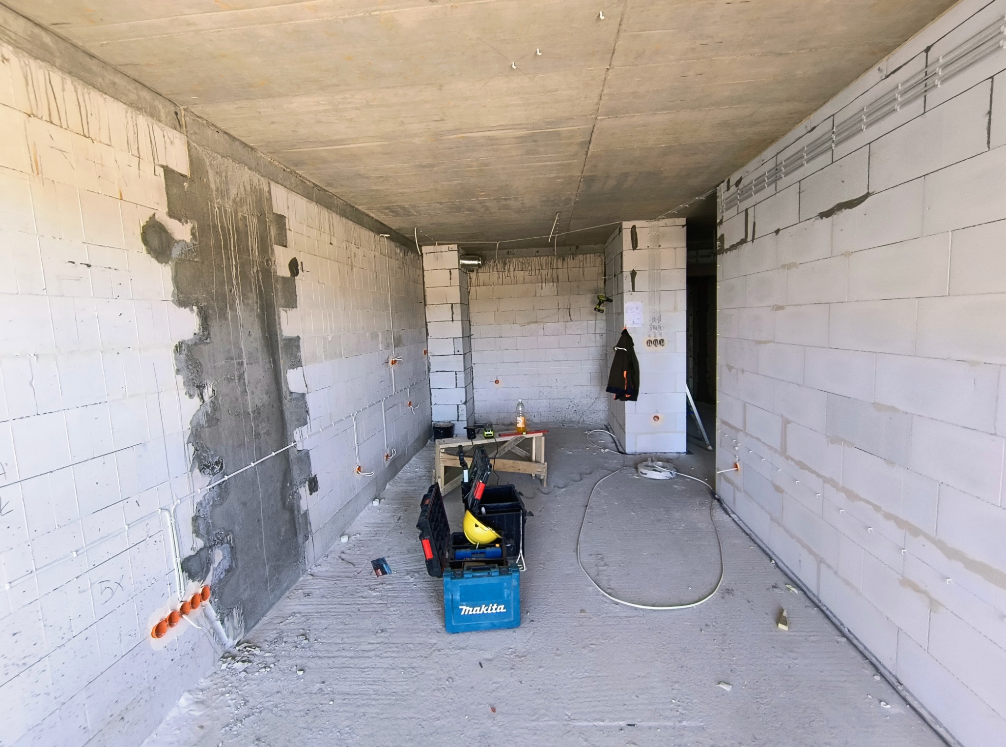 Surowy stan wnętrza z widocznymi elementami instalacji elektrycznej: kable w peszlach, puszki podtynkowe w ścianie z betonu komórkowego, skrzynka narzędziowa Makita i żółty kask na podłodze.