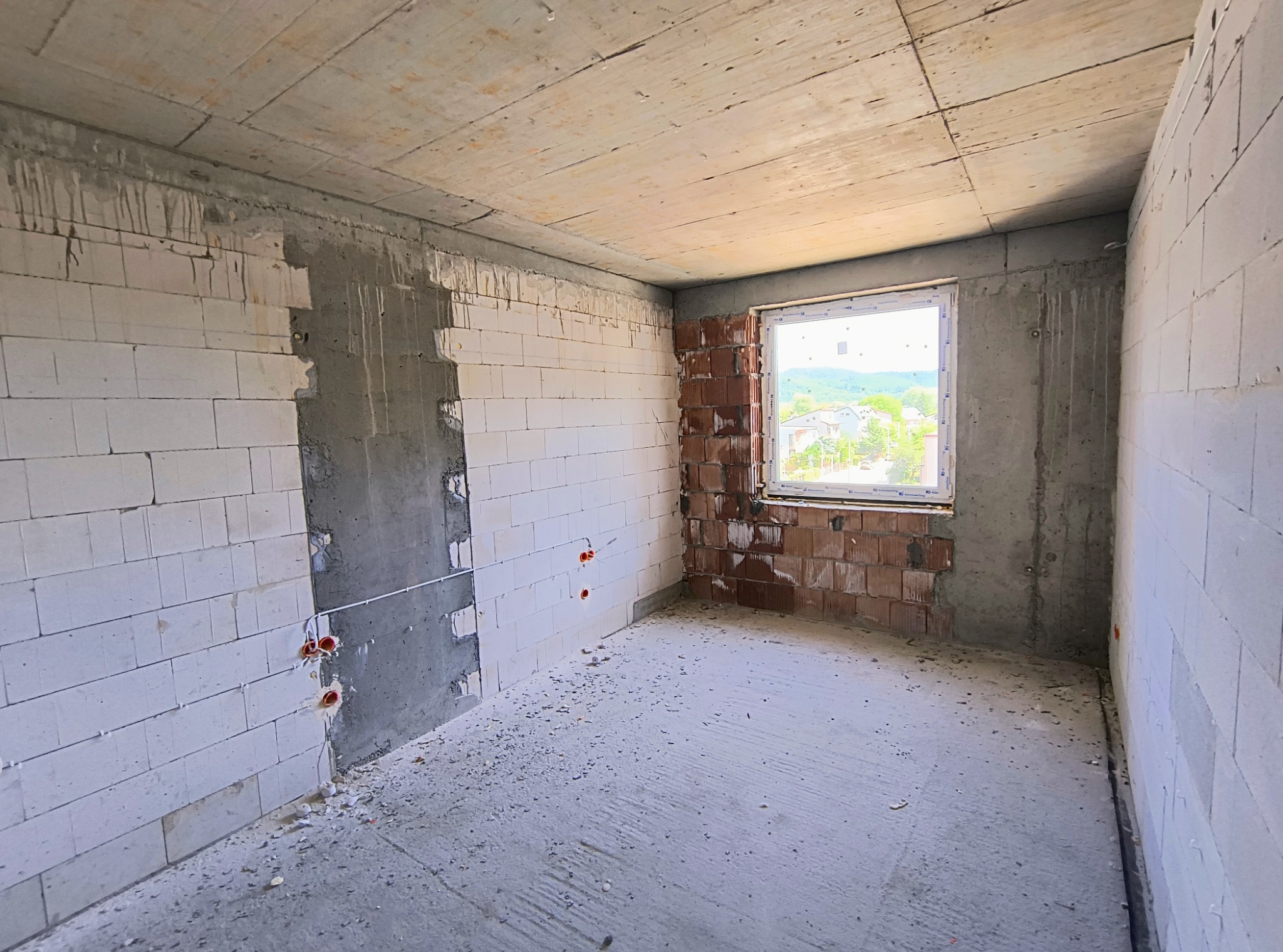 Surowe wnętrze w stanie deweloperskim: ściany z białych bloczków, częściowo otynkowane, z widocznymi puszkami instalacyjnymi; okno z widokiem na zabudowę jednorodzinną i zieleń.