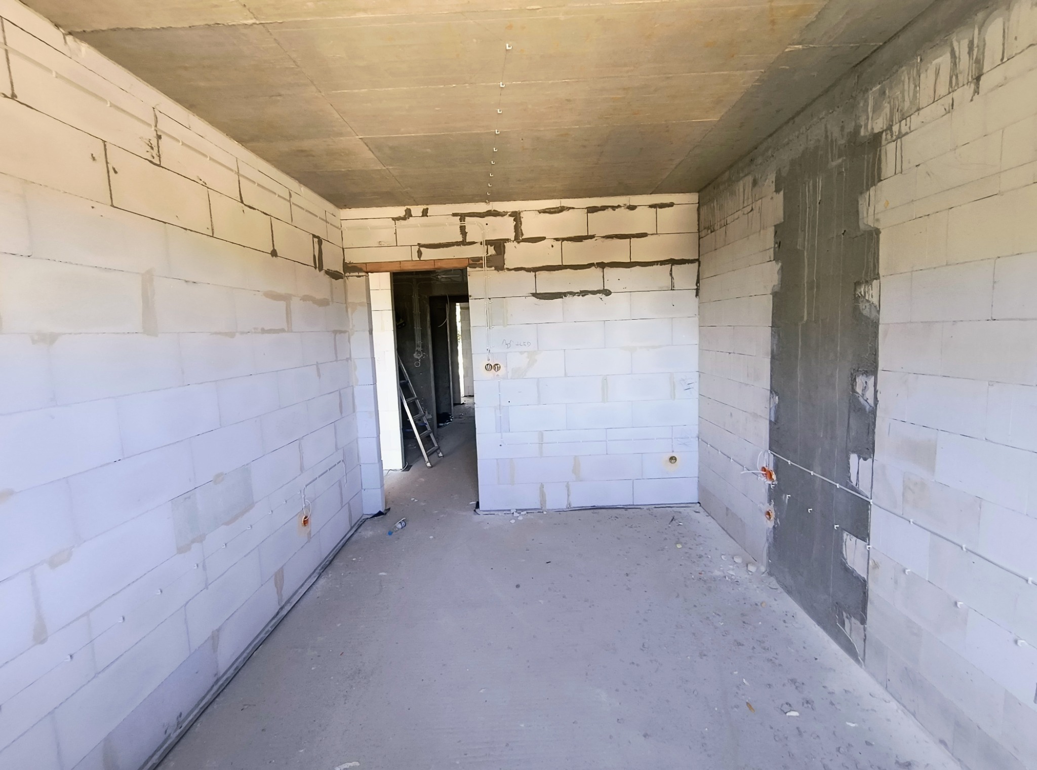 Surowe wnętrze w trakcie budowy, ściany z białych bloczków, widoczne przewody elektryczne i puszki instalacyjne, betonowa podłoga i sufit, drabina w przejściu do kolejnego pomieszczenia.