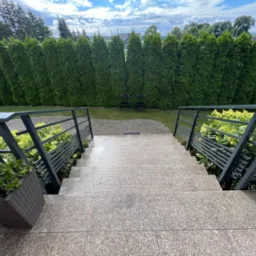 Zewnętrzne schody z szarego granitu z metalową balustradą, donica z roślinami po lewej stronie, w tle wysokie tuje i dwa krzesła.