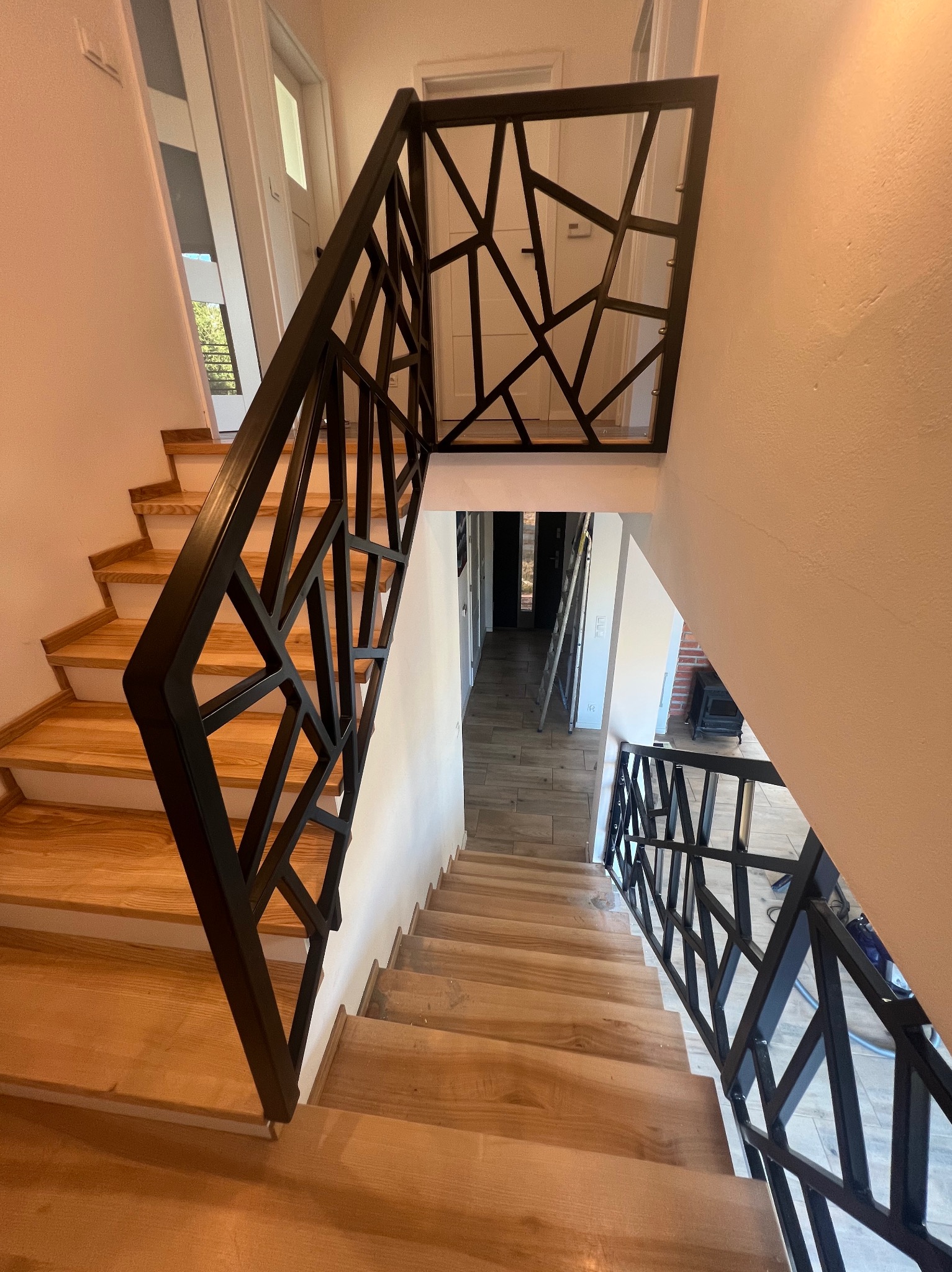 Nowoczesne schody drewniane z czarną, geometryczną balustradą metalową, widok z góry, w tle drabina oparta o ścianę.