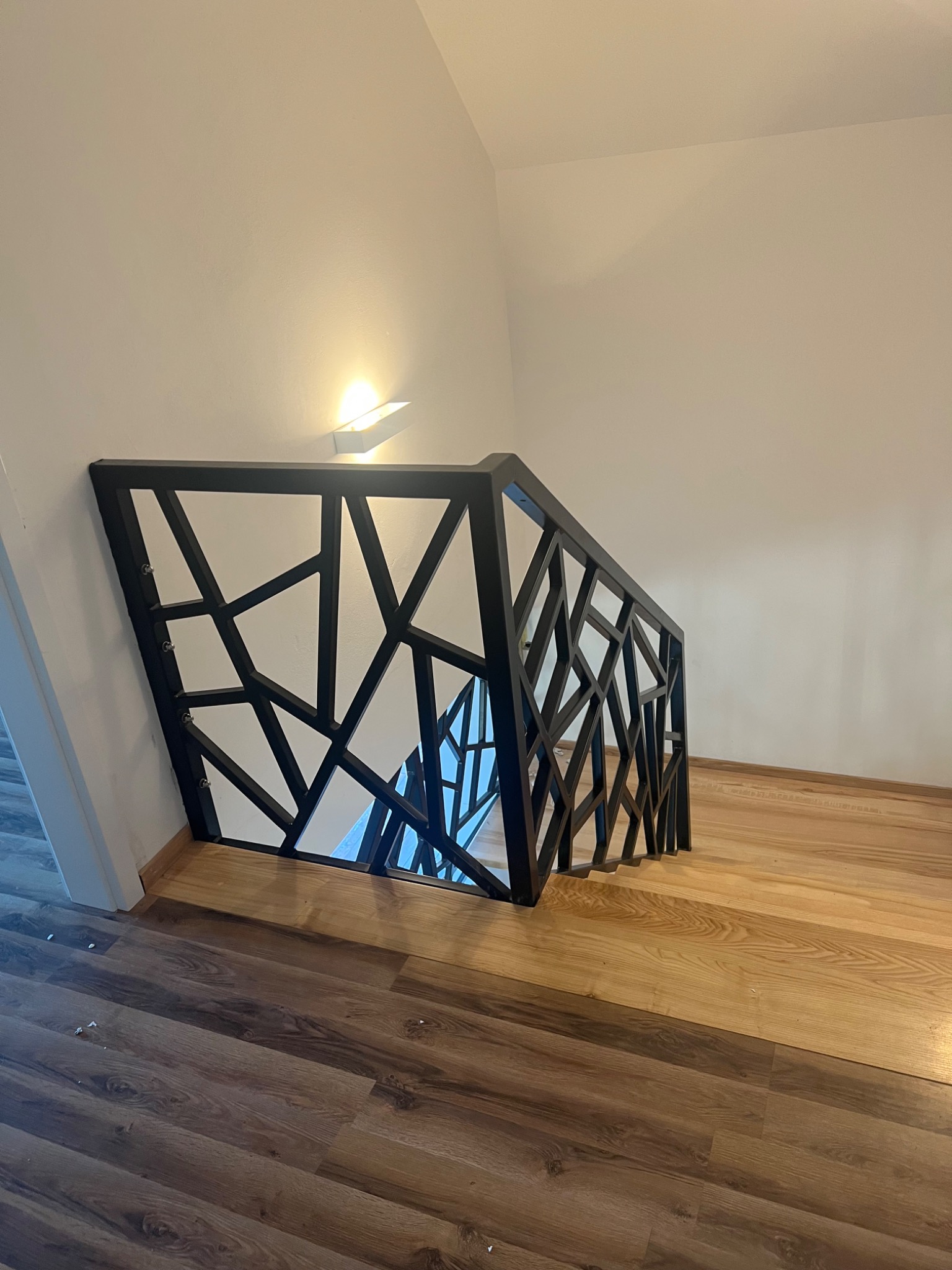 Nowoczesna balustrada schodowa z czarnych, geometrycznych elementów na drewnianych schodach, oświetlona kinkietem.