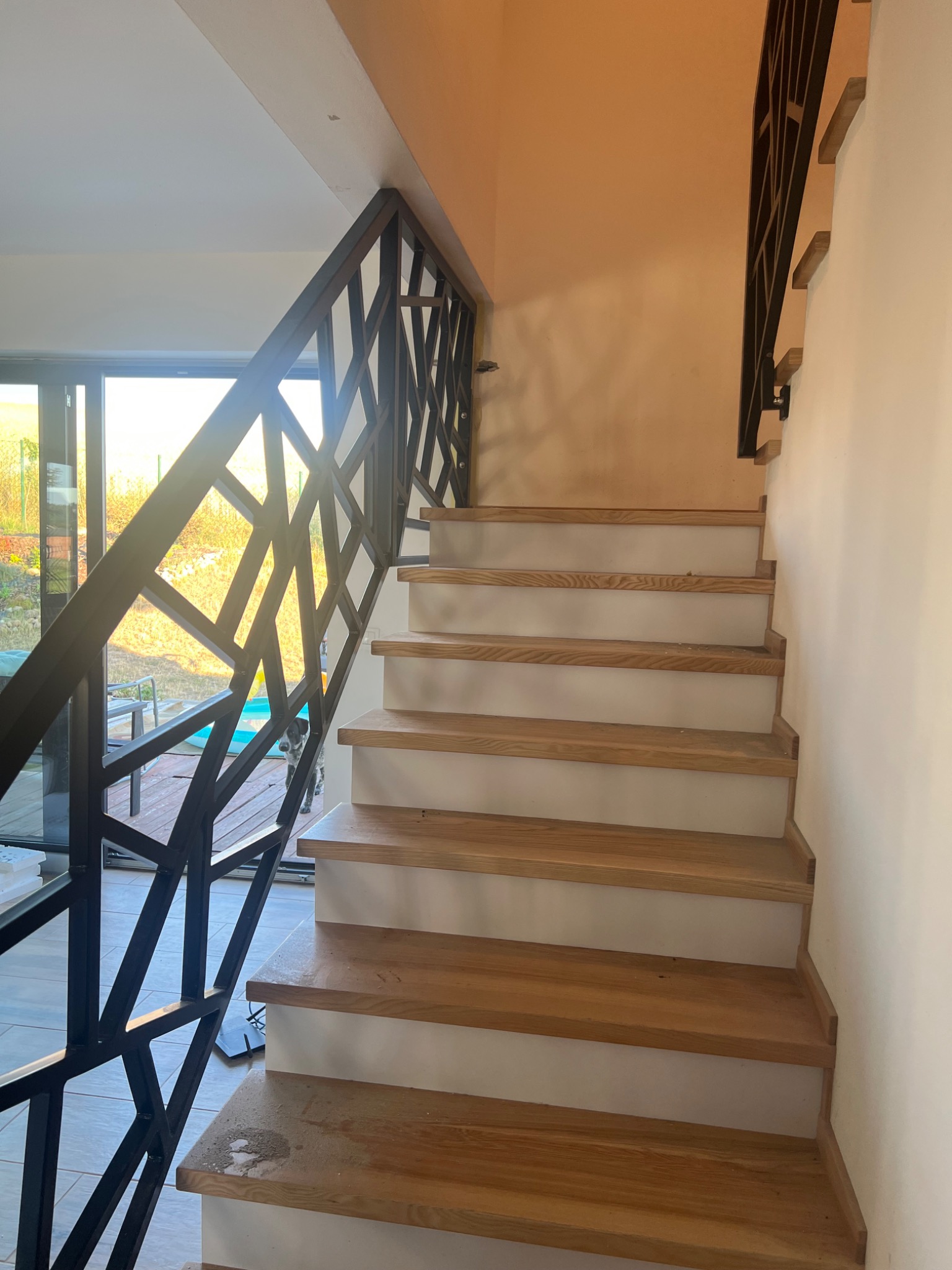Schody drewniane z metalową balustradą o geometrycznym wzorze, widok od dołu, częściowo widoczne otoczenie zewnętrzne przez przeszklone drzwi.