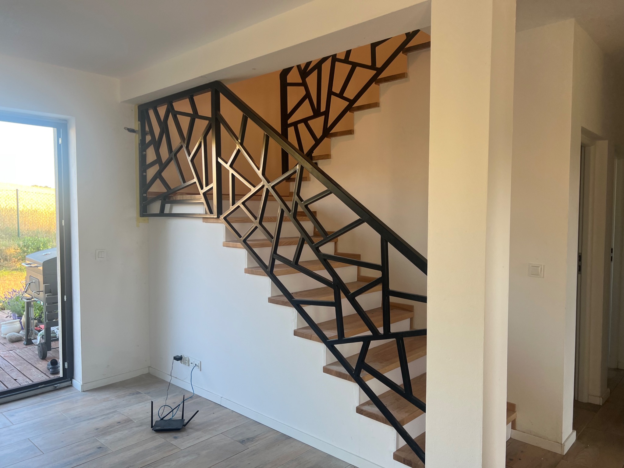 Nowoczesne schody drewniane z czarną, geometryczną balustradą metalową, widok z dołu na jasne wnętrze domu z oknem na zewnątrz.