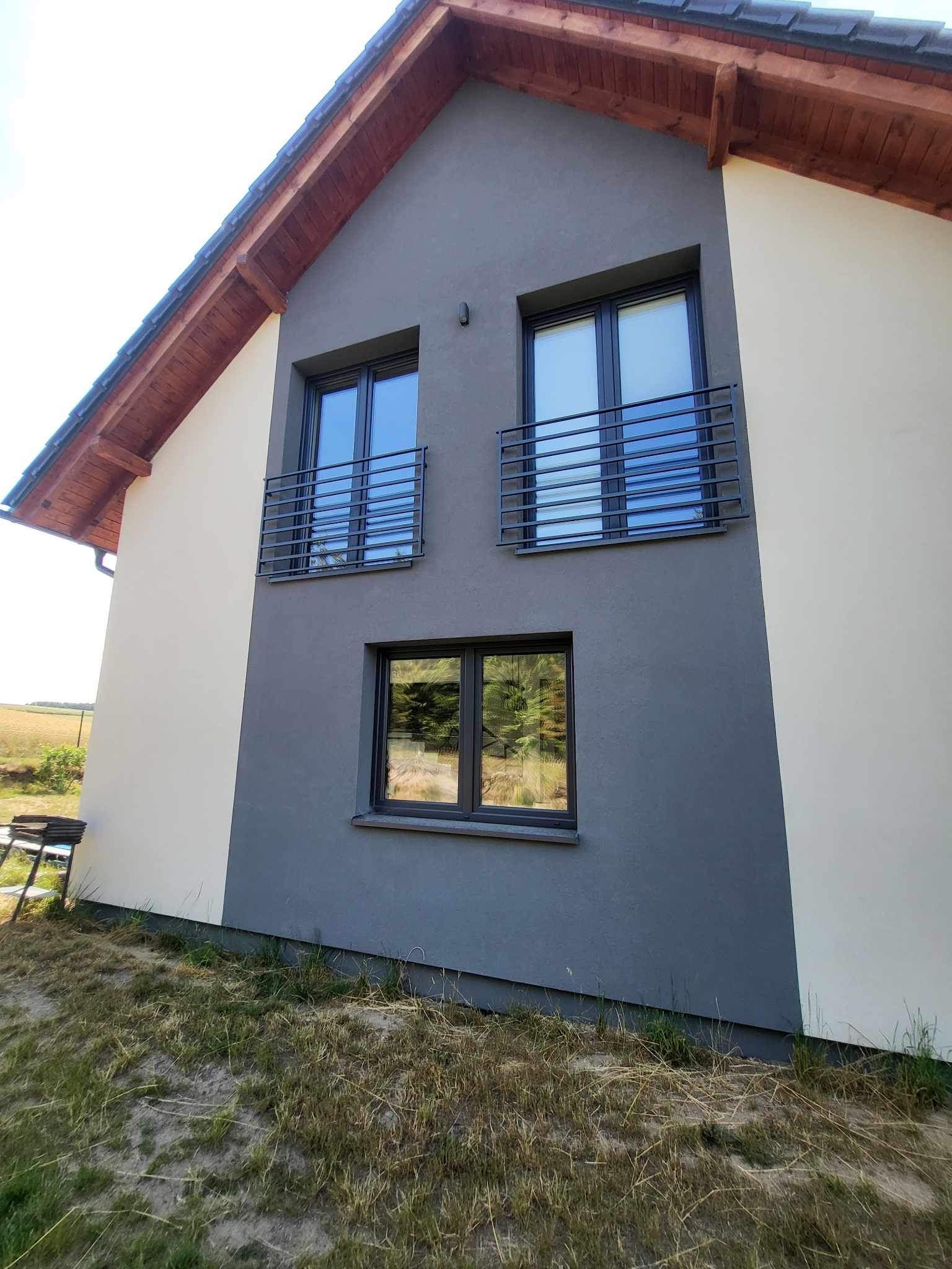 Dwukondygnacyjny dom z szarą elewacją i jasnymi wstawkami, okna z czarnymi ramami i metalowymi balustradami na górnych oknach, widok z boku, trawnik z suchą trawą.