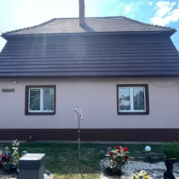 Dom jednorodzinny z nowym, brązowym dachem z dachówki ceramicznej, widok z ogrodu z dekoracjami i wiatraczkiem.