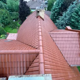 Widok z góry na dach pokryty czerwoną dachówką ceramiczną, z widocznym kominem z cegły i zielonym otoczeniem drzew