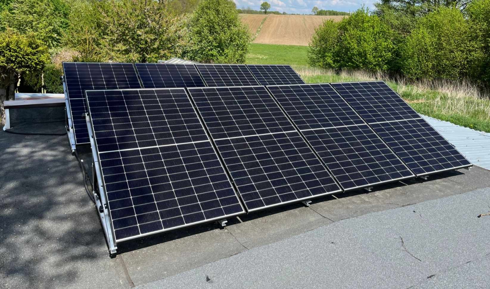 Rząd paneli słonecznych na płaskim dachu z widokiem na zielone drzewa i pole w oddali.
