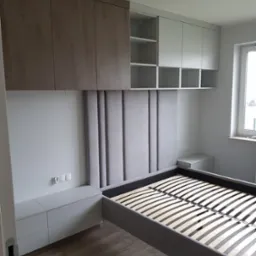 Sypialnia z łóżkiem z szarym tapicerowanym wezgłowiem, szafkami nocnymi i szafkami wiszącymi w kolorze drewna i bieli.