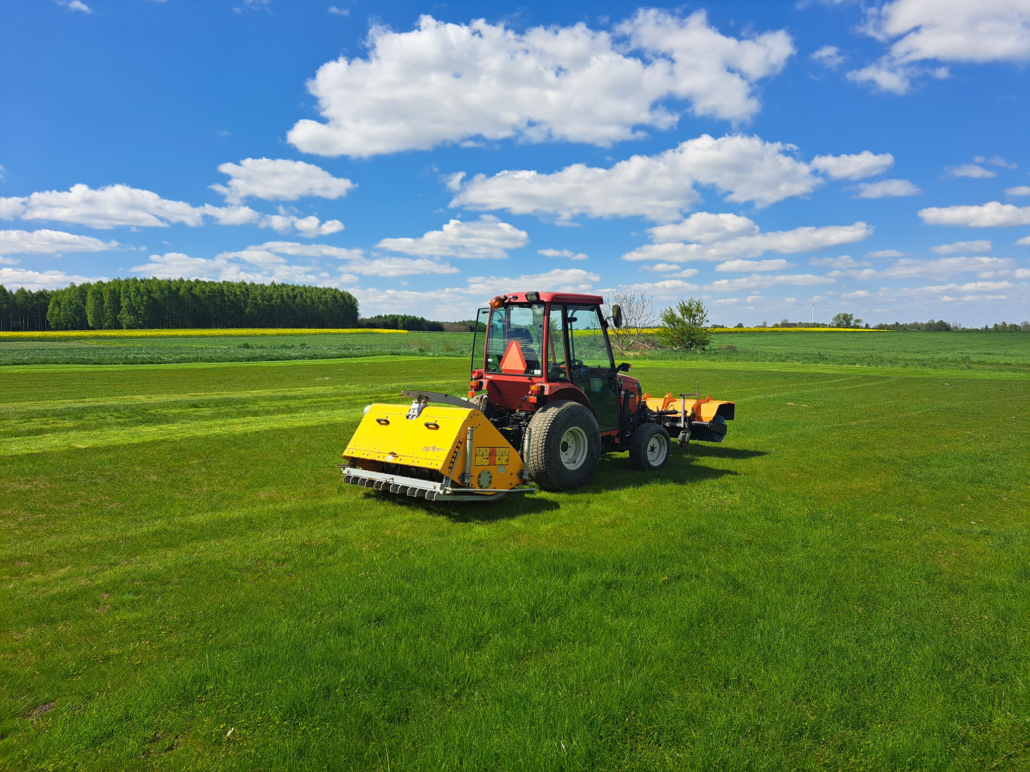 Czerwony traktor z żółtą kosiarką rotacyjną na rozległym, świeżo skoszonym trawniku w słoneczny dzień z błękitnym niebem i białymi chmurami, w tle widoczny pas żółtych kwiatów.