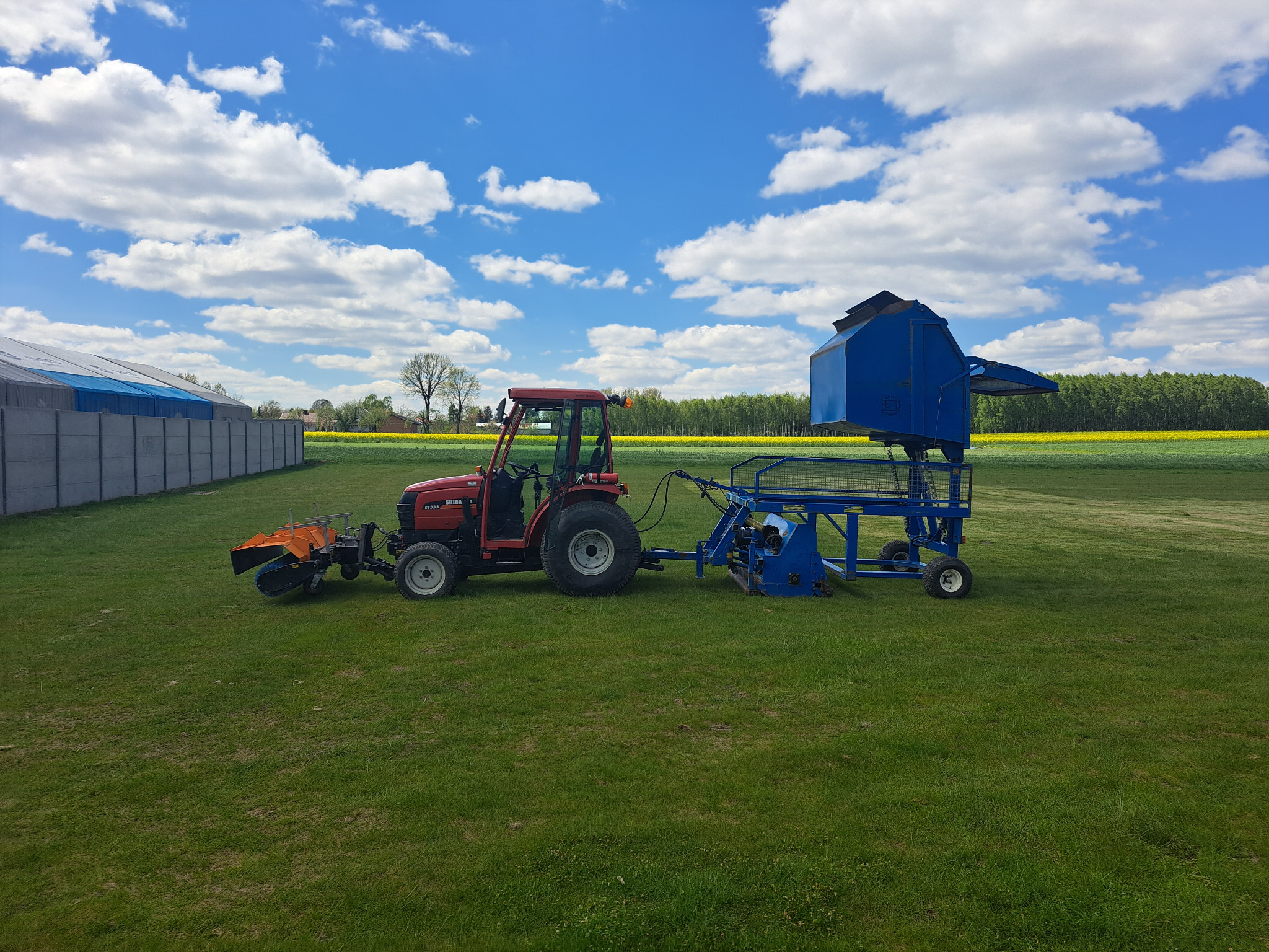 Czerwony traktor Shibaura ST333 z niebieskim osprzętem do zbierania trawy na zielonym polu, w tle widoczne drzewa i błękitne niebo z chmurami.