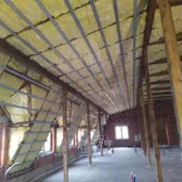 Poddasze w trakcie ocieplania wełną mineralną, widoczne drewniane belki konstrukcyjne, metalowe profile, okna w murze z czerwonej cegły i instalacje sanitarne na podłodze.