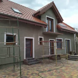 Dom w trakcie nakładania szarego tynku zewnętrznego, okna i drzwi zabezpieczone folią, widoczne rusztowania i kostka brukowa na podwórku.
