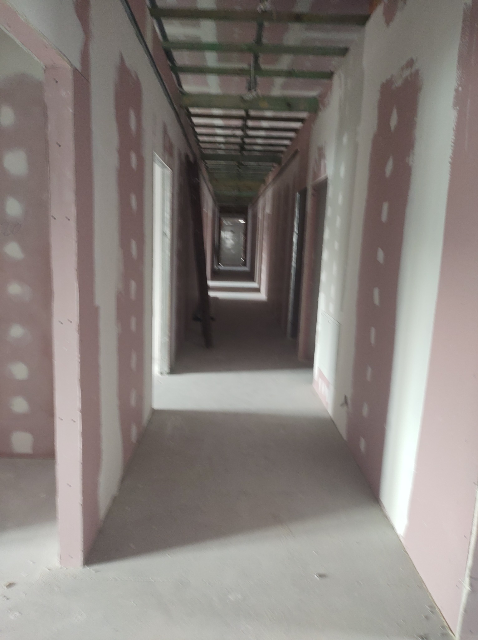 Długi korytarz w trakcie remontu, ściany wykończone płytami gipsowo-kartonowymi, widoczne elementy konstrukcji sufitu, przygotowania do dalszych prac wykończeniowych.