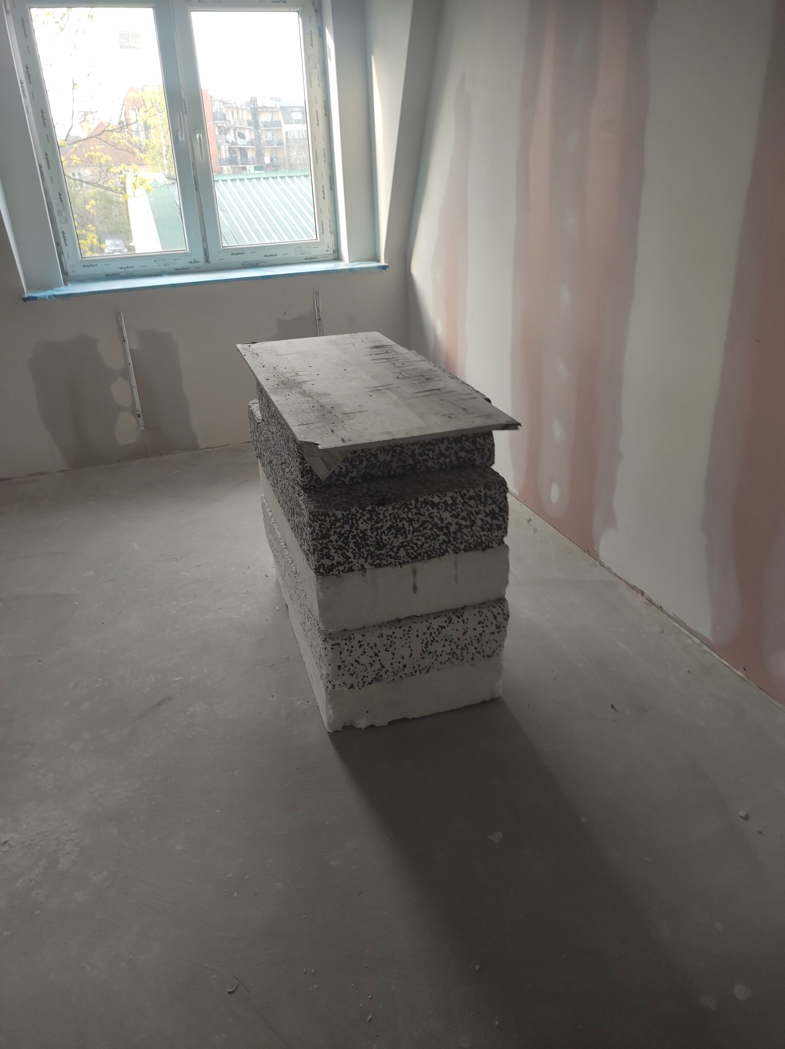 Styropianowe płyty izolacyjne ułożone w stos na szarym podłożu w pomieszczeniu w trakcie prac wykończeniowych, widoczne ślady gładzi na ścianach i okno na poddaszu.