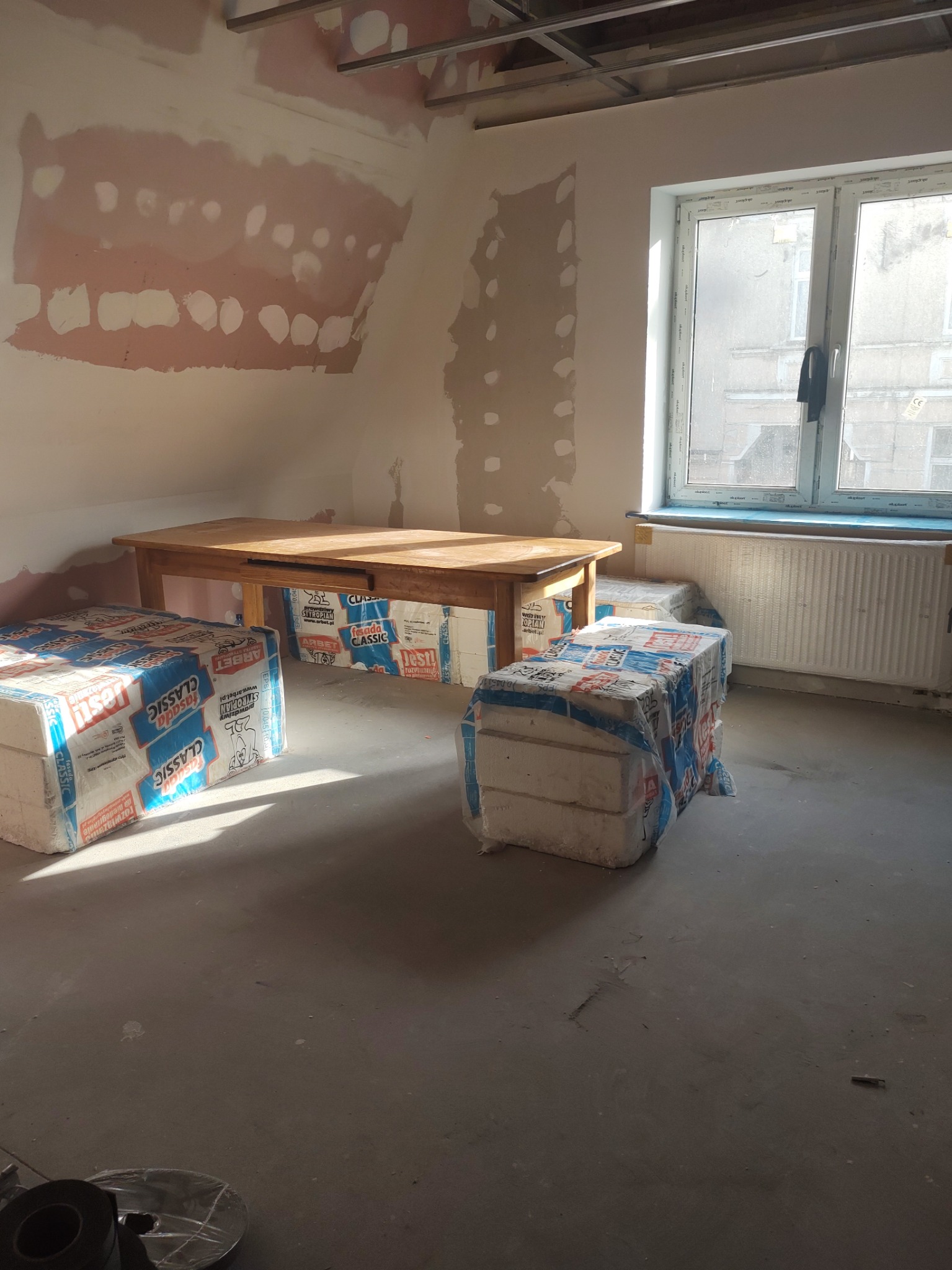 Wnętrze w trakcie remontu: ściany z widocznymi śladami po wcześniejszych pracach, drewniany stół, paczki styropianu pod stołem i obok, okno z grzejnikiem pod parapetem.