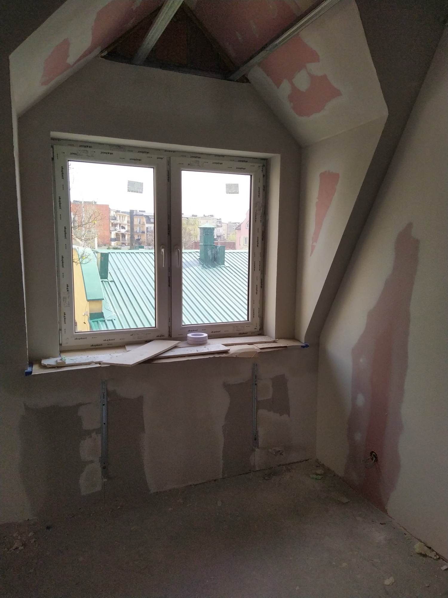 Poddasze w trakcie remontu: widoczne okno z widokiem na zielony dach, ściany z fragmentami gładzi gipsowej i metalowy stelaż sufitu
