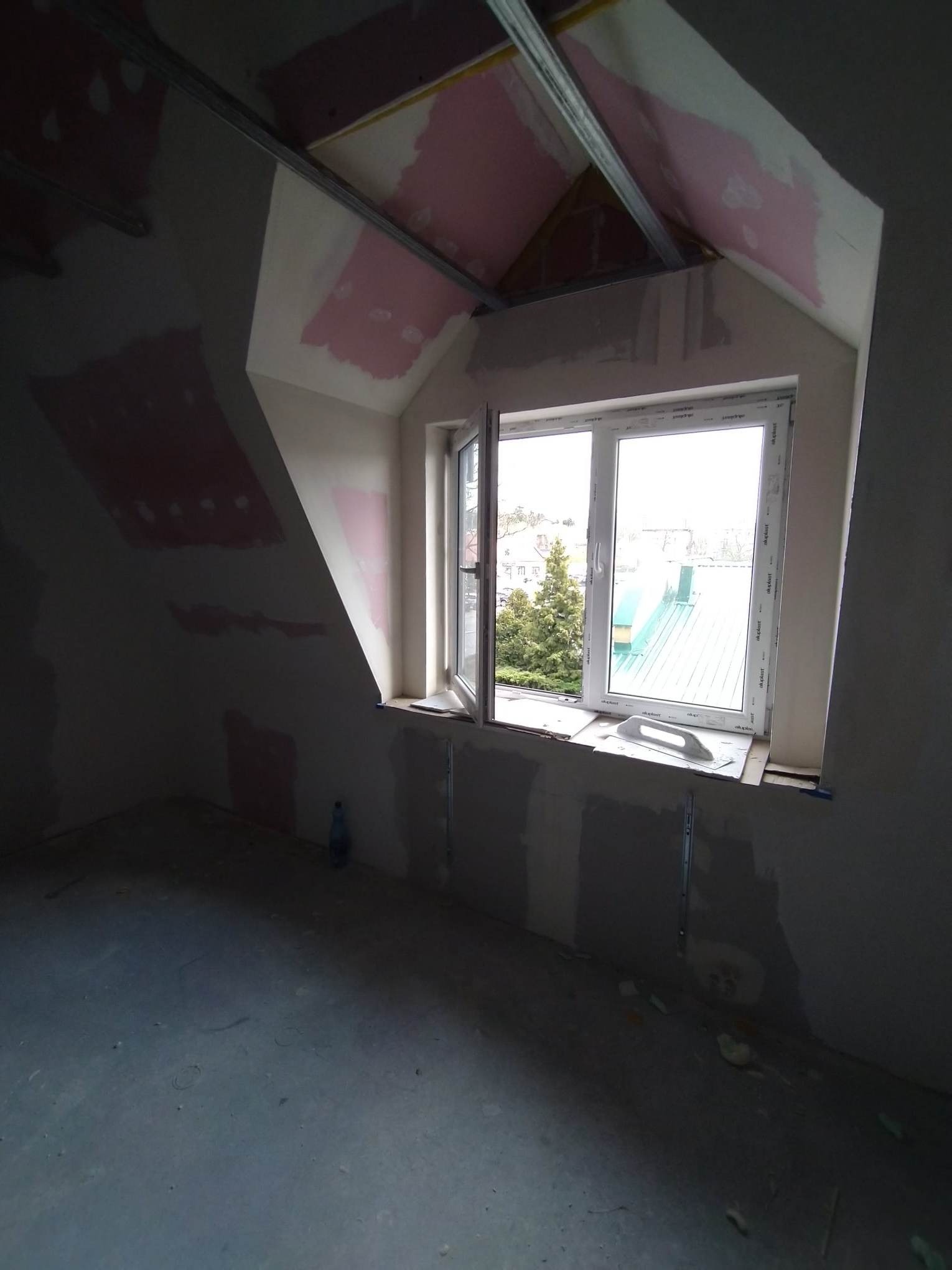 Poddasze w trakcie adaptacji: widok na okno z widocznymi elementami konstrukcji, częściowo zagruntowane ściany i sufit, oraz metalowe profile.