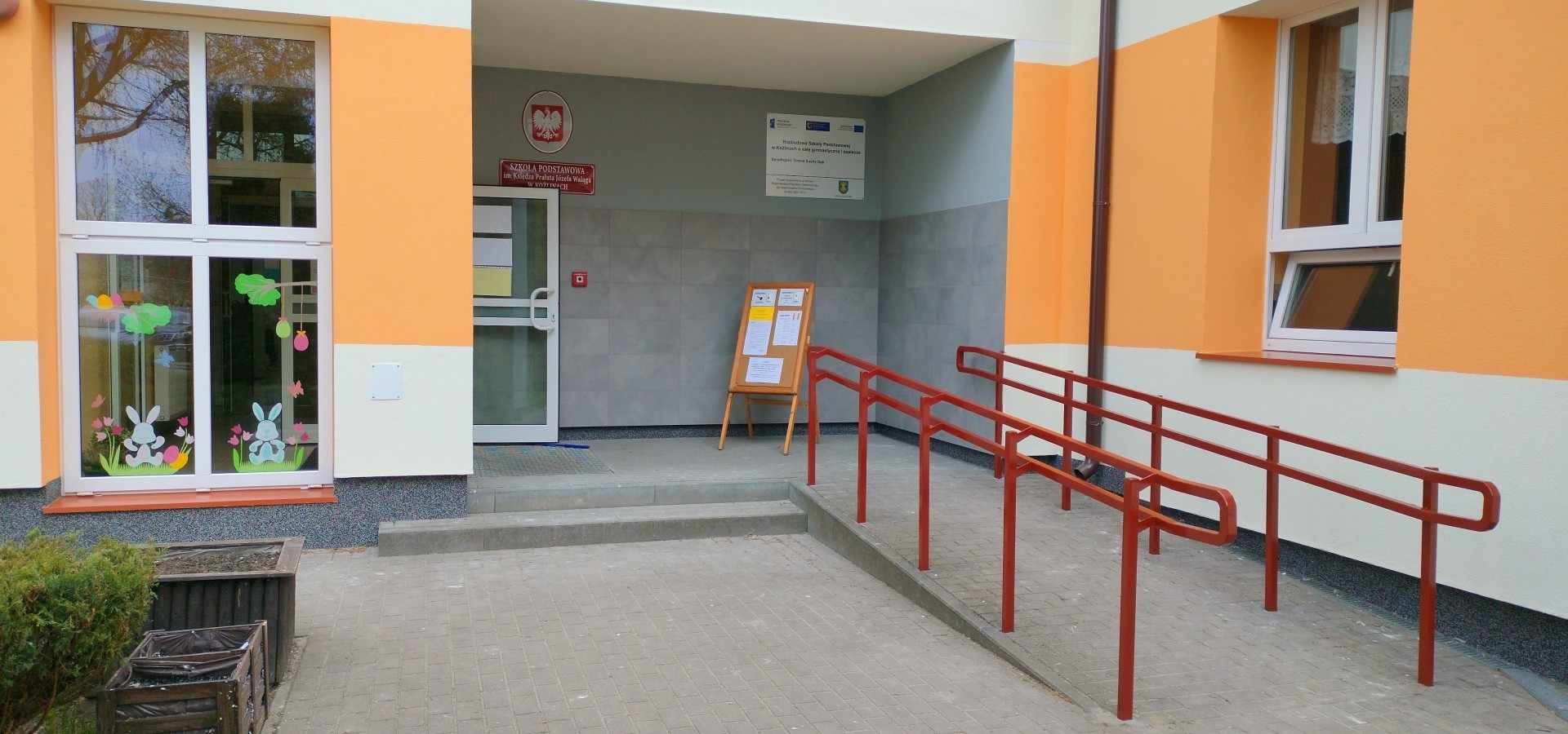 Wejście do budynku szkoły podstawowej z nową, pomarańczowo-białą elewacją, rampą dla niepełnosprawnych z czerwonymi poręczami i oknami ozdobionymi naklejkami wielkanocnymi.