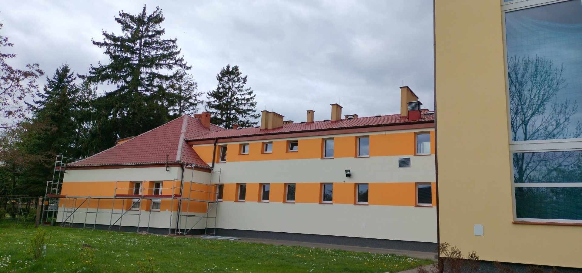 Dwukondygnacyjny budynek z pomarańczowo-kremową elewacją w poziome pasy, czerwony dach, widoczne rusztowania przy bocznej ścianie, na tle drzew i pochmurnego nieba.
