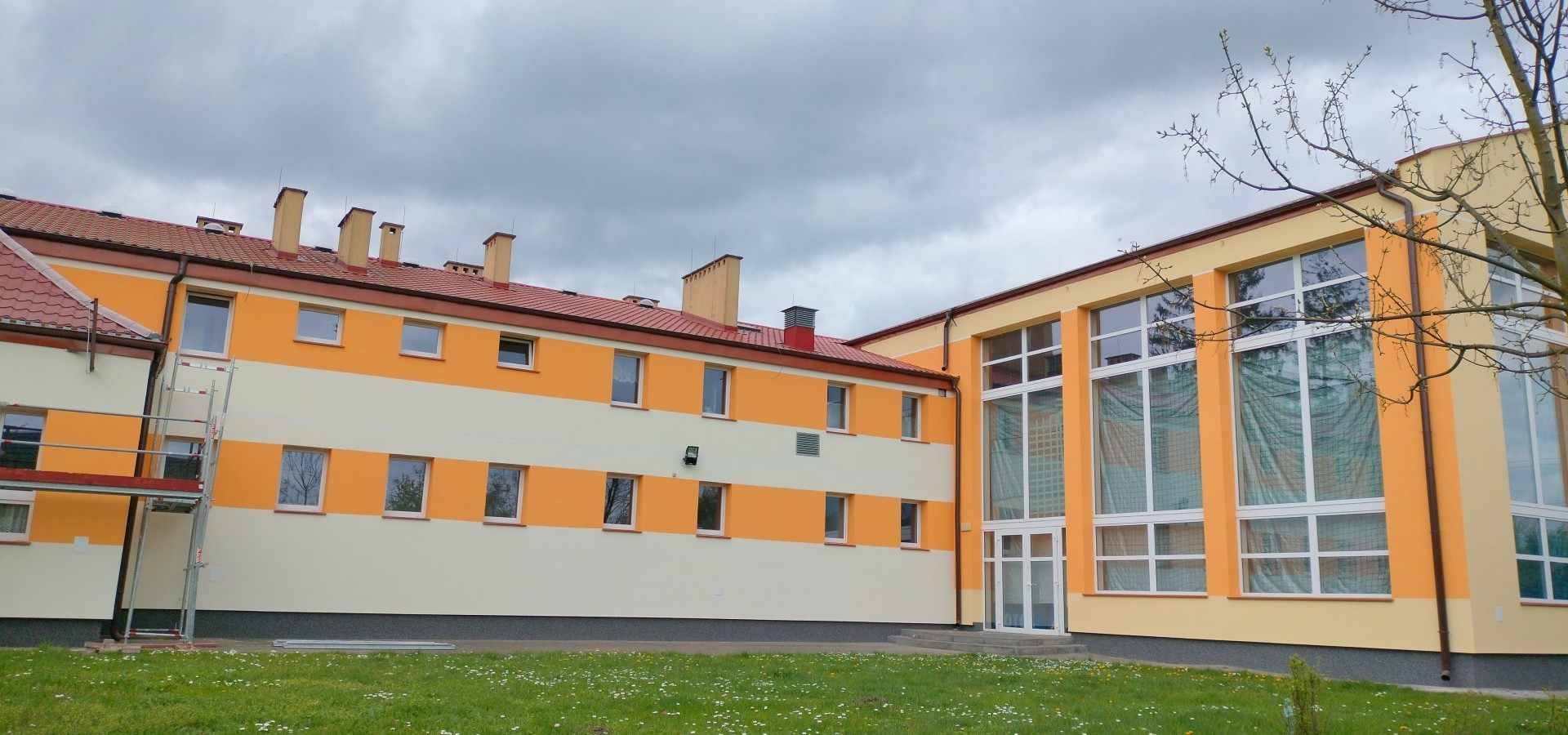 Dwukondygnacyjny budynek z nową, pomarańczowo-kremową elewacją, widziany z poziomu trawnika z kwitnącymi stokrotkami; rusztowanie przy bocznej ścianie, szare niebo.