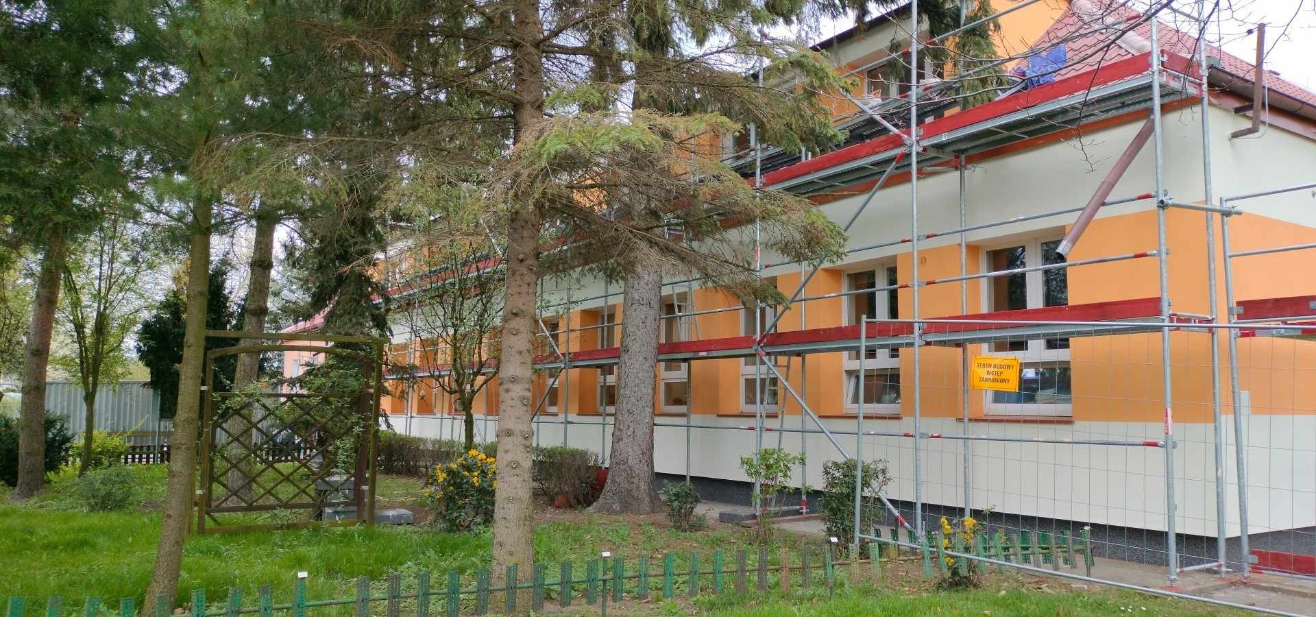 Budynek wielorodzinny w trakcie prac elewacyjnych, widoczne rusztowania, żółta tablica informacyjna 'teren budowy wstęp zabroniony', drzewa na pierwszym planie.