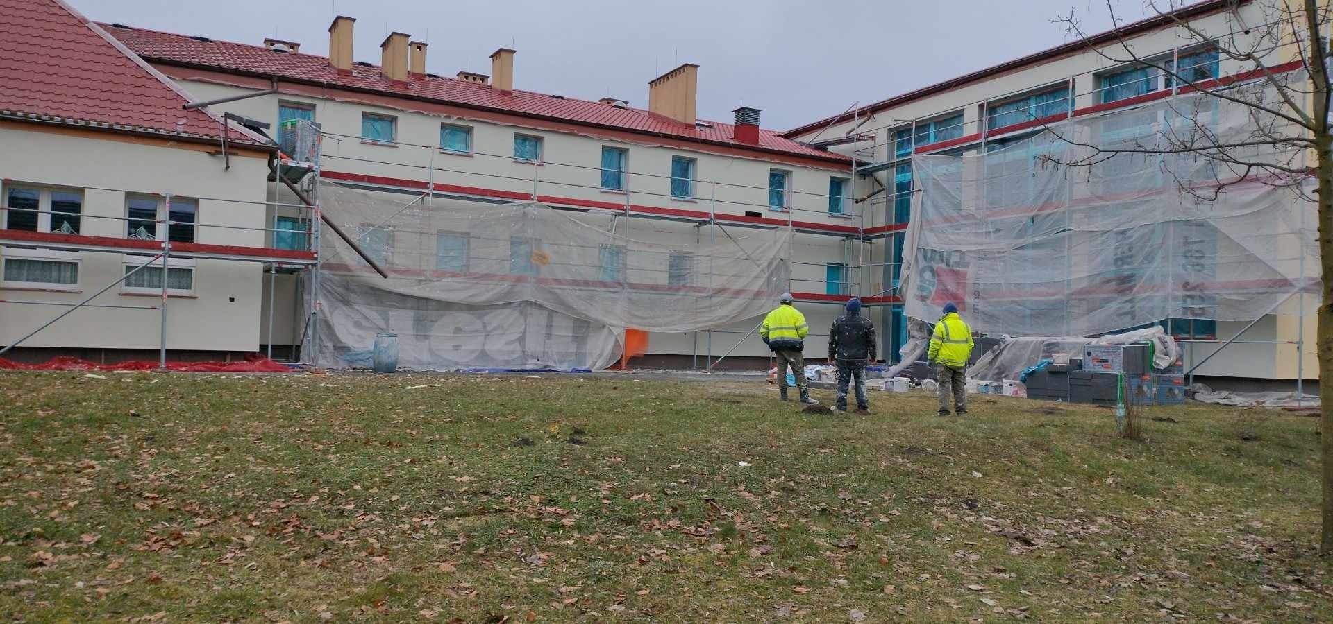 Trzech pracowników w odblaskowych kamizelkach stoi na trawniku przed budynkiem w trakcie prac elewacyjnych, rusztowania i siatka ochronna zasłaniają fragment fasady, widoczne okna zabezpieczone folią.