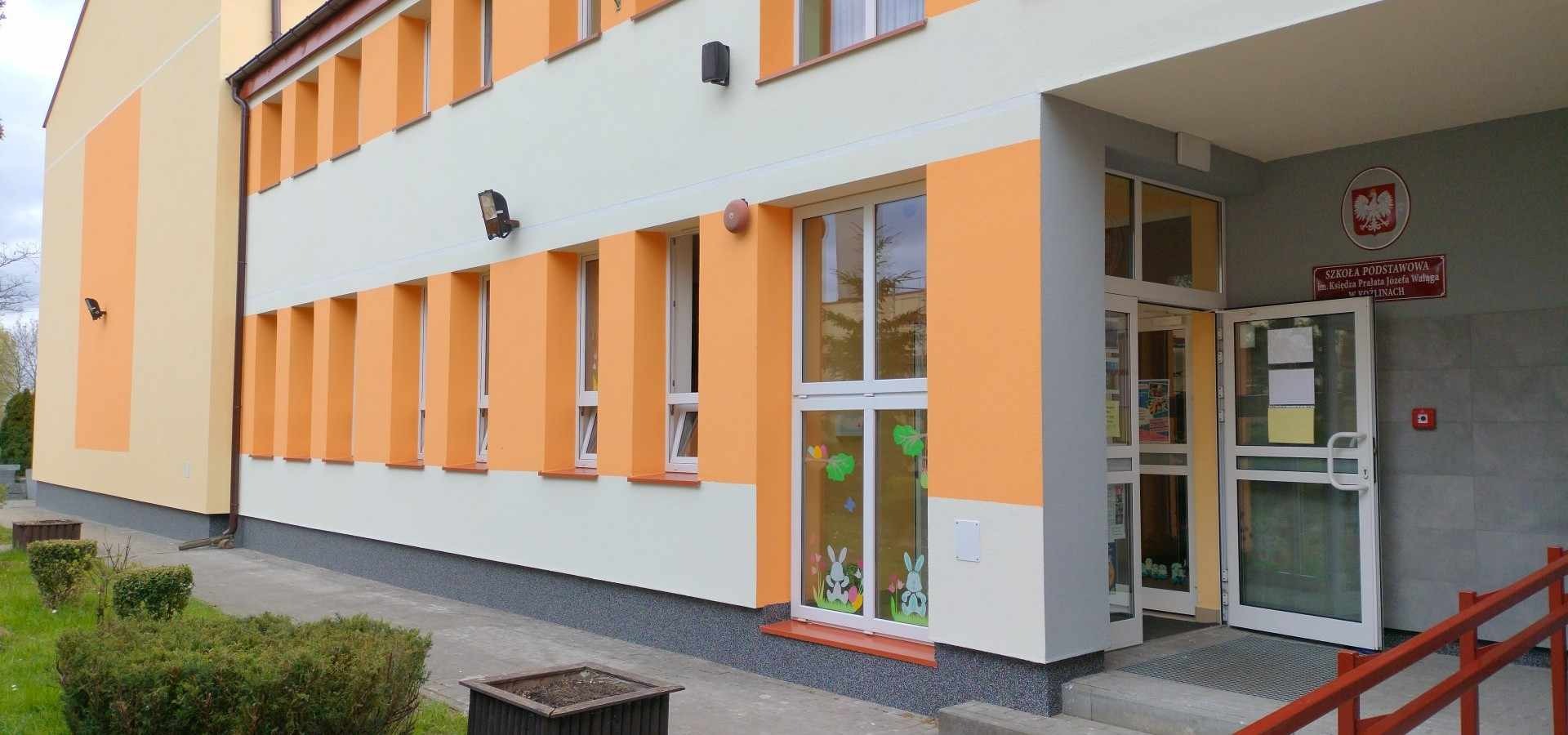 Odnowiona elewacja budynku szkoły podstawowej z pomarańczowymi i białymi elementami, okna ozdobione naklejkami z motywem królików, widoczne wejście główne z czerwonymi barierkami.