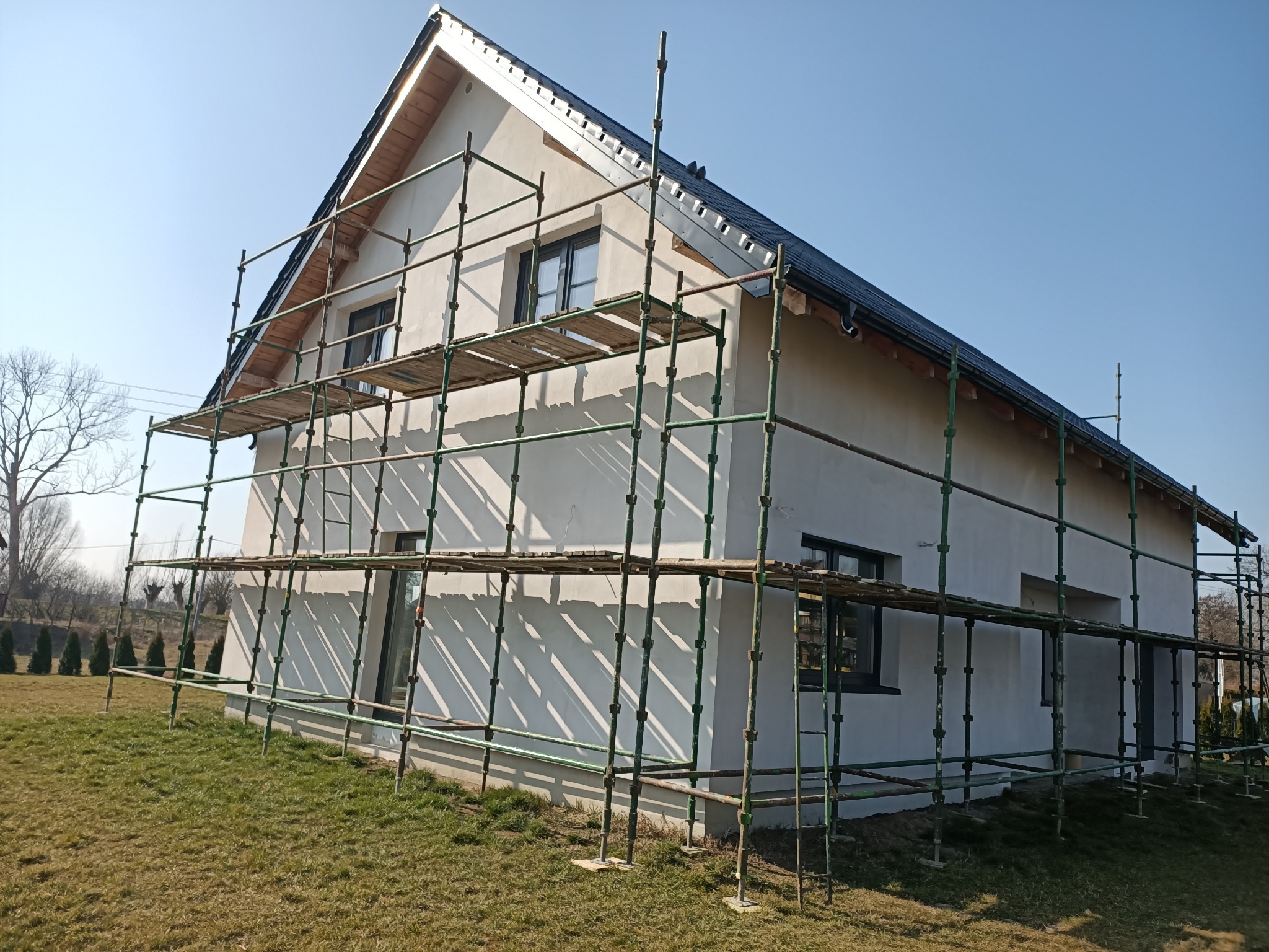 Dom jednorodzinny w trakcie ocieplania elewacji, widoczne rusztowania i świeżo nałożony tynk w jasnym odcieniu. Dach pokryty ciemną dachówką.