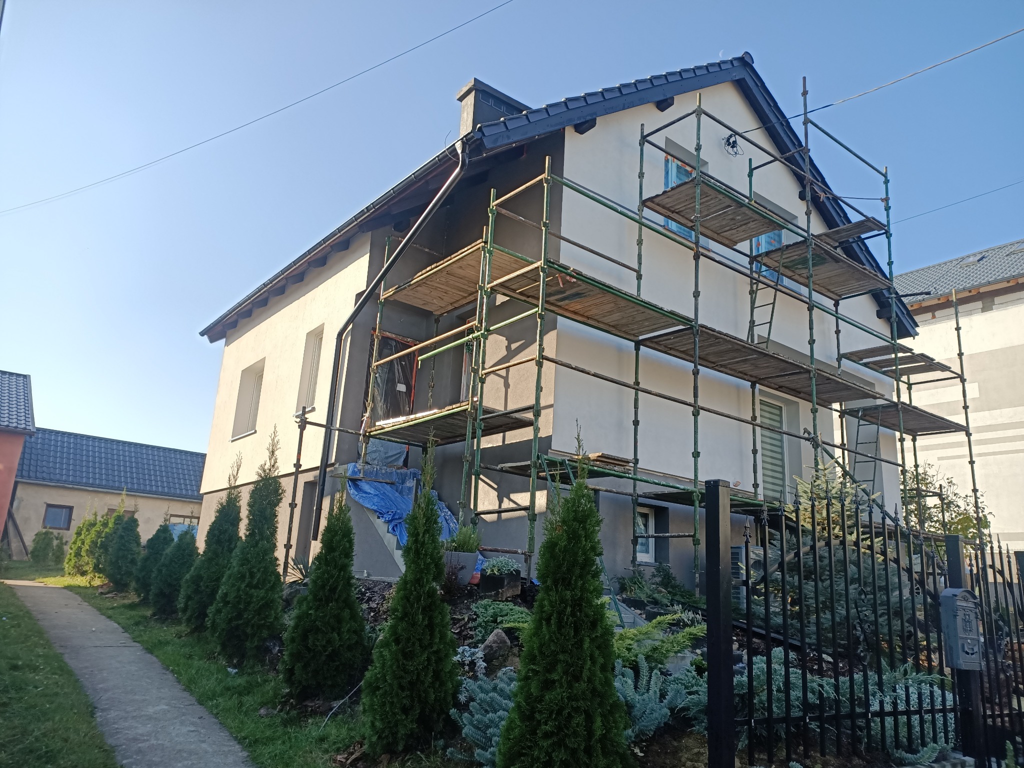 Dwukondygnacyjny dom jednorodzinny w trakcie prac elewacyjnych, z ustawionymi rusztowaniami i częściowo otynkowaną fasadą w odcieniach szarości i bieli, widok z poziomu ogrodu z tujami.