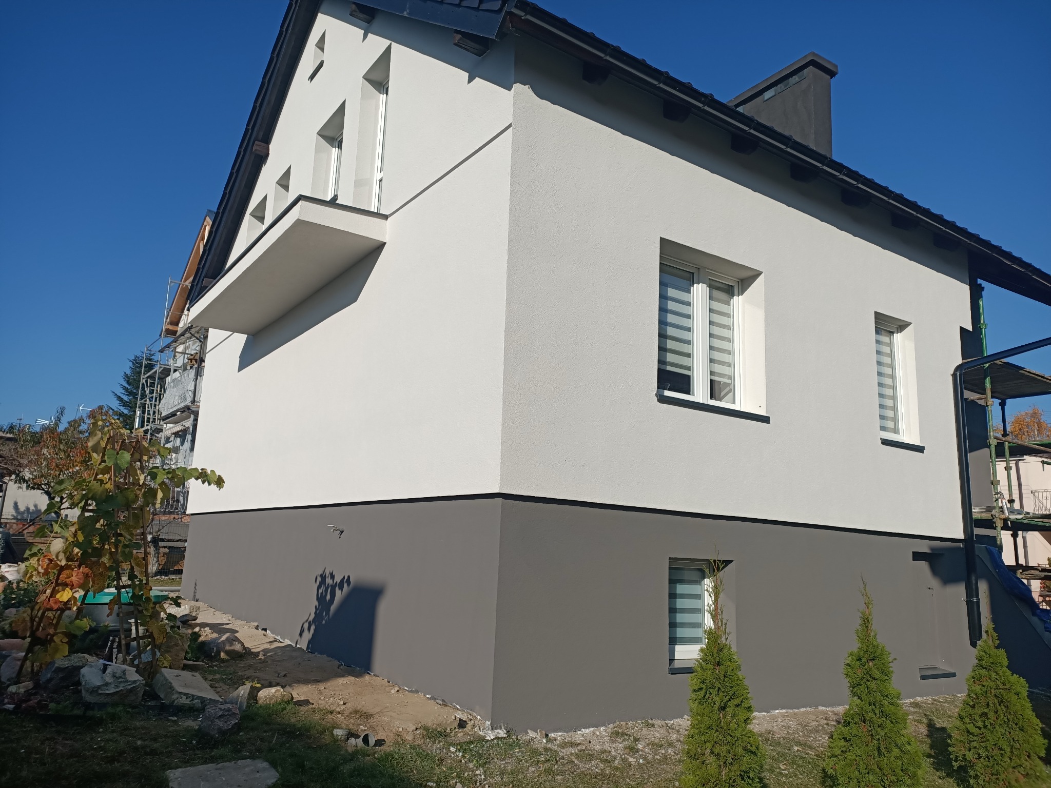 Dwukondygnacyjny dom z wykończoną elewacją w odcieniach szarości i bieli, widoczne rusztowania z boku budynku, małe drzewka iglaste przy cokole i winorośl przy ścianie.