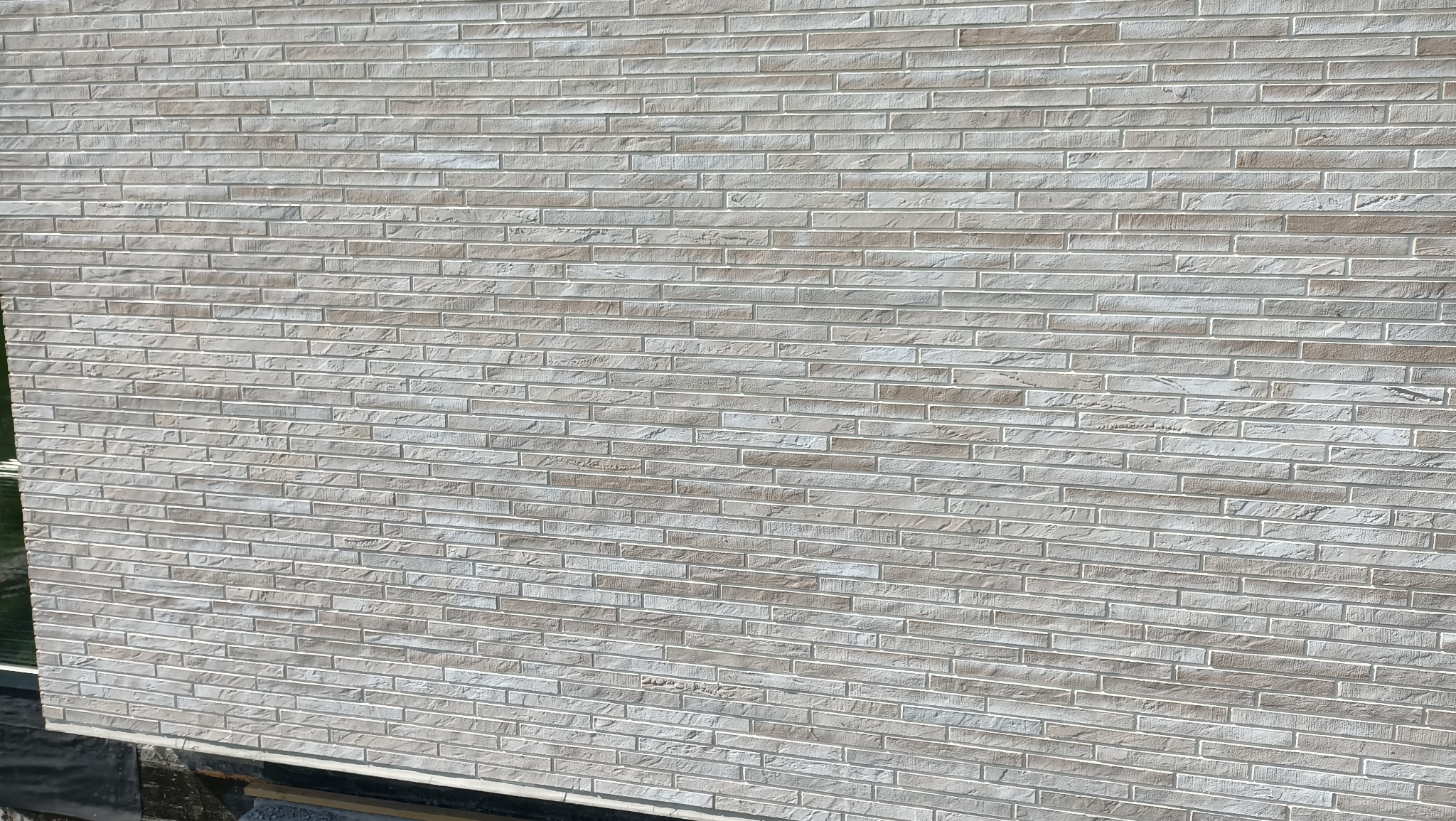 Elewacja budynku wykończona jasną, beżową cegłą klinkierową z wyraźną fugą. Widoczny fragment okna z lewej strony. Ujęcie z dołu ukazujące strukturę i kolorystykę elewacji.