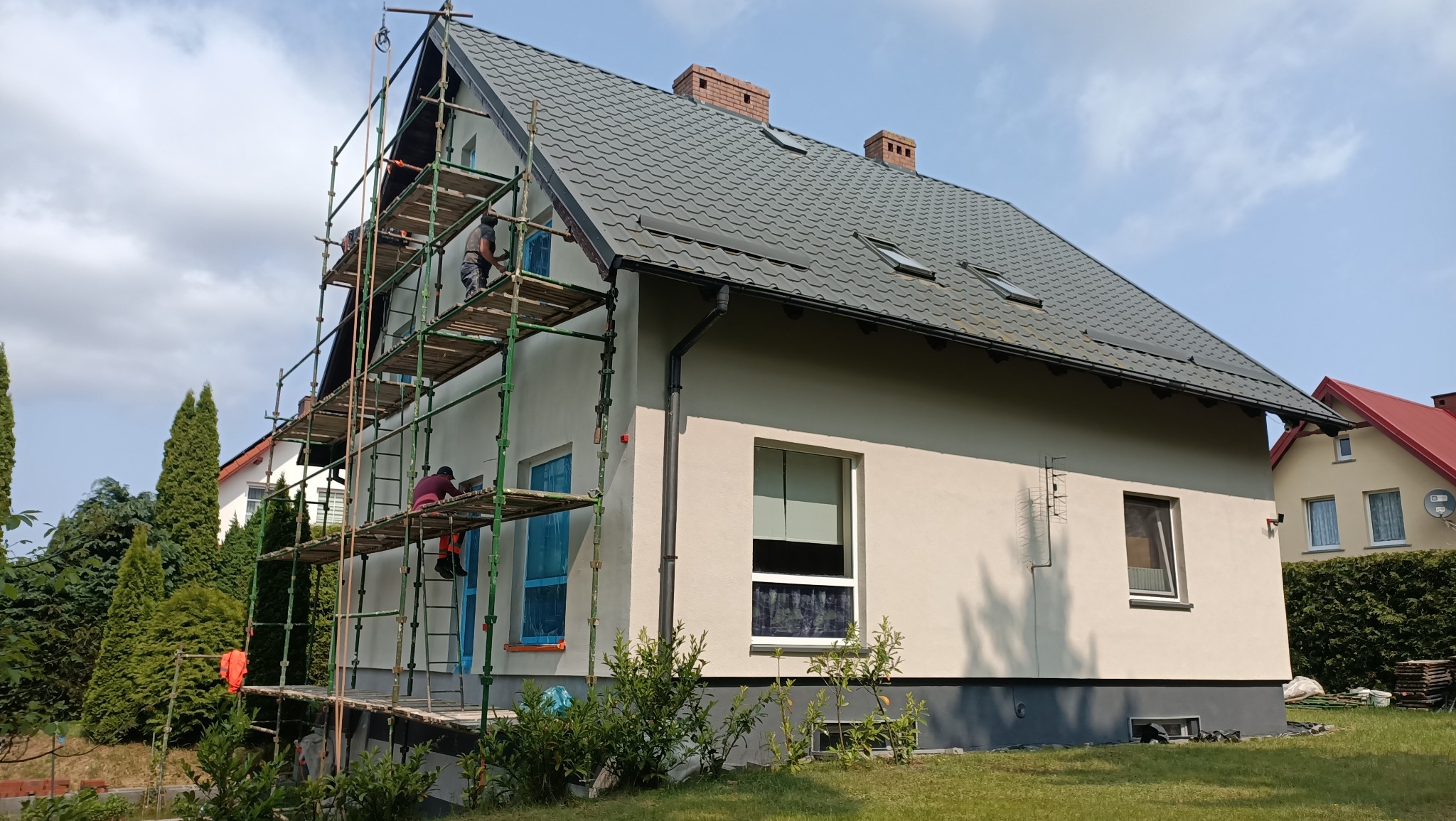Dom jednorodzinny po renowacji elewacji, z pracownikami na rusztowaniu. Okna zabezpieczone folią, widoczna świeżo nałożona warstwa tynku.