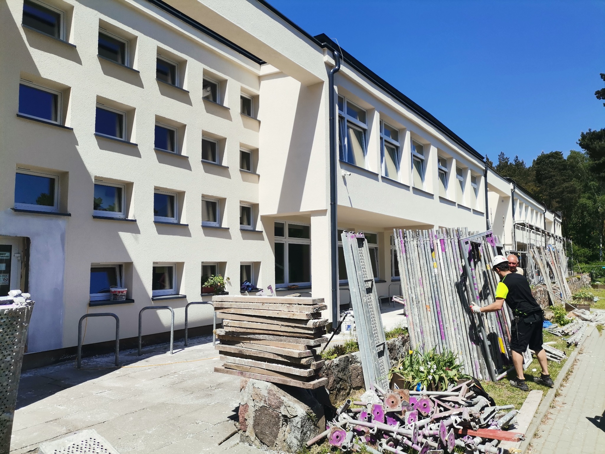 Pracownik w kasku ustawia rusztowanie przy budynku z jasną elewacją, widoczne deski, narzędzia i elementy rusztowania na ziemi.