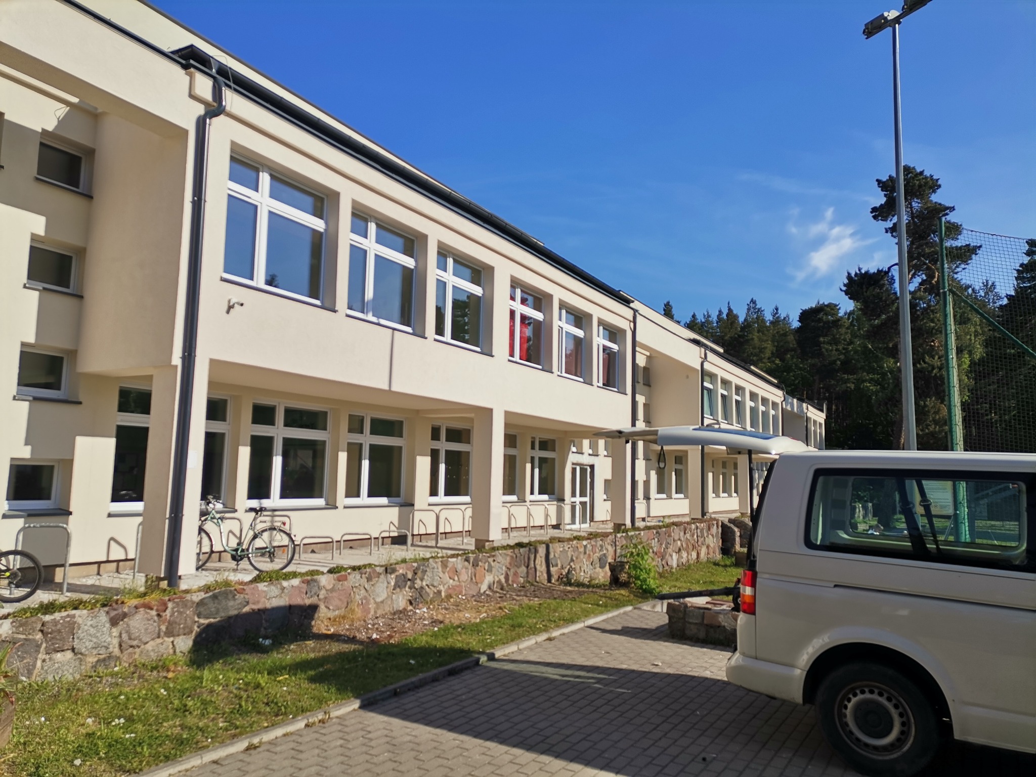 Budynek po ociepleniu elewacji, widoczny z boku z zaparkowanym białym busem i rowerami przy stojaku, jasna elewacja, białe okna, błękitne niebo.