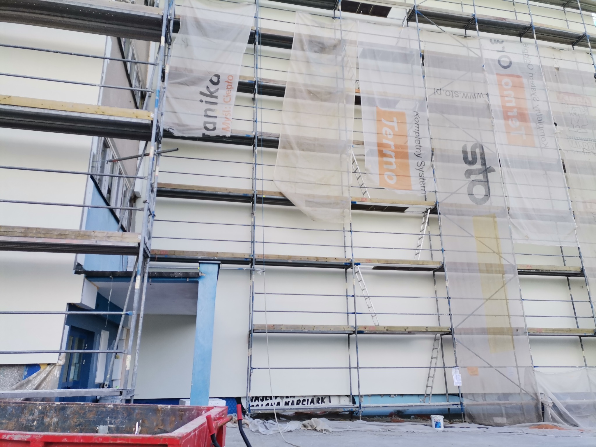 Budynek w trakcie prac elewacyjnych, widoczne rusztowania z siatkami reklamowymi firm Termo i STO, kontener na odpady budowlane na pierwszym planie.