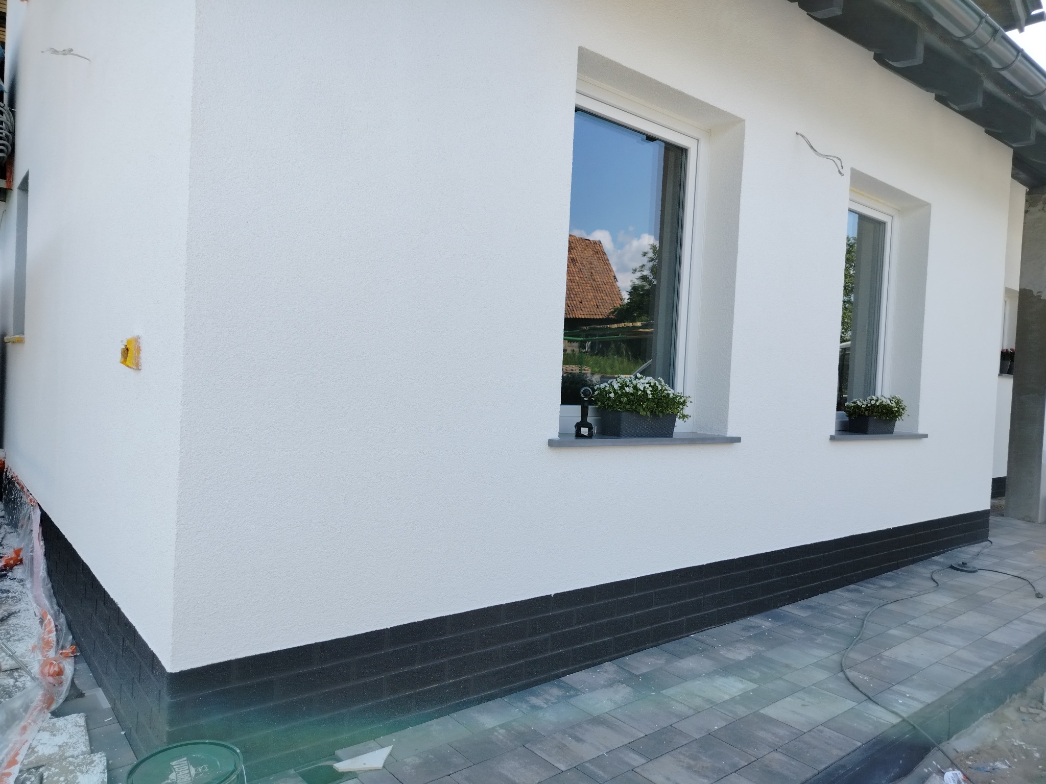 Biała elewacja domu jednorodzinnego z dwoma oknami, na których stoją doniczki z kwiatami; cokół wykończony ciemną cegłą klinkierową, widoczny fragment chodnika z kostki brukowej.